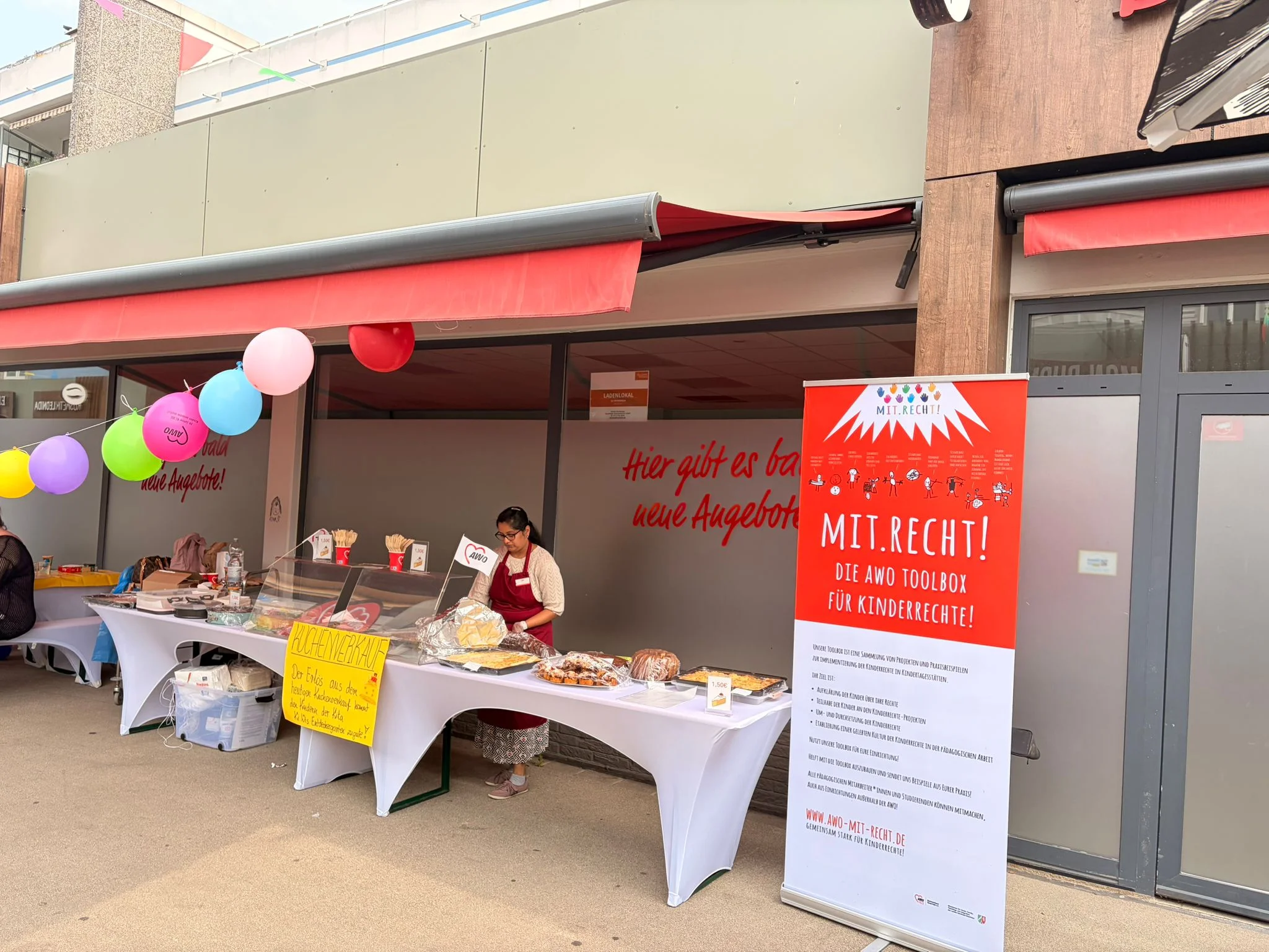 Auf dem Foto ist ein Aktionsstand der AWO vor einem Gebäude zu sehen. Links stehen mehrere mit weißen Tischhussen verkleidete Tische mit Kuchen, Gebäck und weiteren Speisen sowie einem handgeschriebenen Schild „Kuchenverkauf“, darüber sind bunte Luftballons aufgehängt. Rechts im Bild steht ein großer rot-weißer Aufsteller mit der Aufschrift „MIT.RECHT! Die AWO Toolbox für Kinderrechte!“.