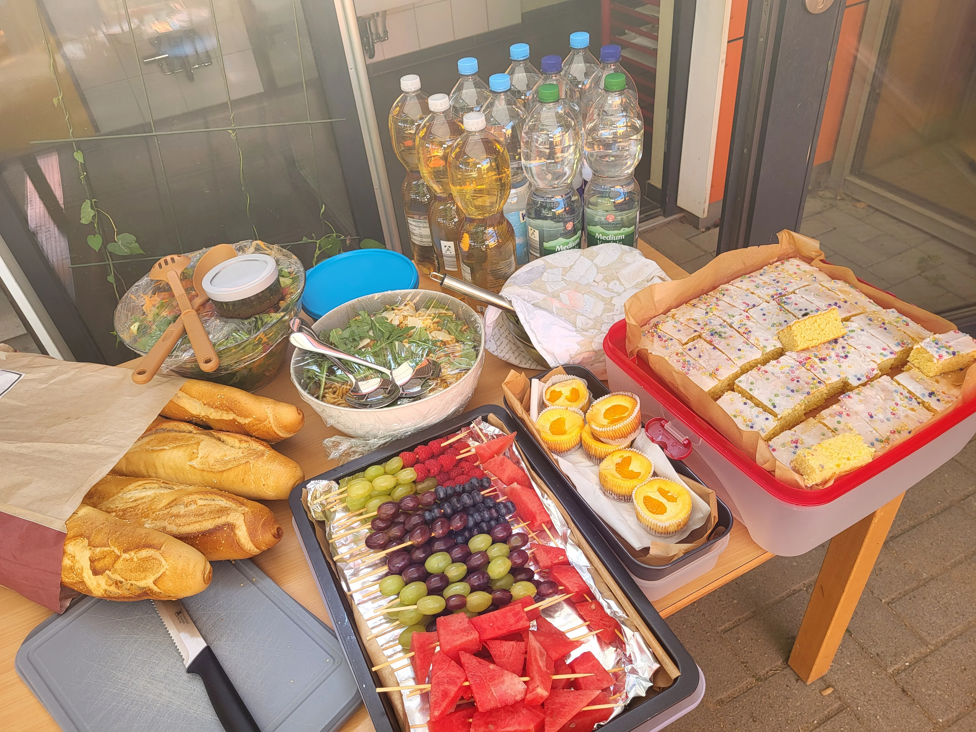 Auf dem Bild ist ein Buffet beziehungsweise eine kleine Tafel mit Essen und Getränken im Außenbereich zu sehen. Auf dem Tisch stehen mehrere Schüsseln mit Salat, belegte Obstspieße (Trauben, Melone, Beeren), daneben Baguettebrote in einer Papiertüte sowie ein Tablett mit Kuchenstücken mit weißem Guss und Streuseln in einer roten Box. Außerdem sind Muffins mit Aprikosenhälften, mehrere Flaschen Wasser und Saft bzw. Schorle im Hintergrund sowie ein Brotmesser und ein Schneidebrett zu erkennen.