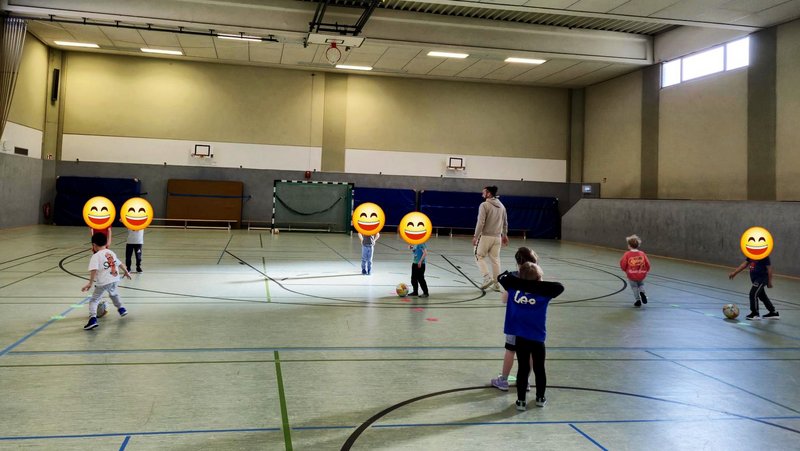 Auf dem Bild sieht man eine Sporthalle, in der mehrere Kinder gemeinsam mit einem Erwachsenen Fußball spielen – die Kinder bewegen sich frei im Raum, einige mit Bällen.