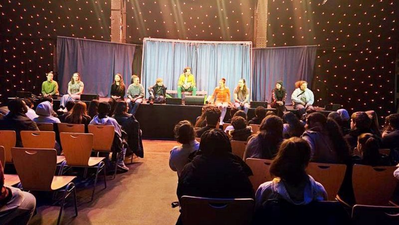 Auf dem Bild ist eine Szene in einem Theater oder Auditorium während einer Veranstaltung zu sehen. Hier sind die Details: Vordergrund und Mittelgrund (Zuschauerraum): Man sieht eine Gruppe von Zuschauern, die auf Holzstühlen sitzen und zur Bühne blicken. Die Köpfe der Zuschauer im Vordergrund verdecken teilweise die Sicht auf die Reihen dahinter. Hintergrund (Bühne): Auf einer erhöhten Bühne sitzt eine Gruppe von ungefähr zehn jungen Menschen in Freizeitkleidung. Sie scheinen an einer Podiumsdiskussion, einer Fragerunde oder einer Präsentation teilzunehmen. In der Mitte der Gruppe sitzt eine Person in einer leuchtend gelben Jacke. Hinter der Gruppe ist ein hellblauer/türkiser Vorhang. Beleuchtung und Atmosphäre: Der Raum ist dunkel gehalten, aber die Bühne ist durch mehrere helle Scheinwerfer von oben stark beleuchtet. Die Hintergrundwand seitlich der Bühne ist mit einem dunklen Stoff verkleidet und mit vielen kleinen, leuchtenden Punkten (eventuell Lichterketten oder eine spezielle Beleuchtungsinstallation) versehen, was eine theatralische oder festliche Atmosphäre schafft.