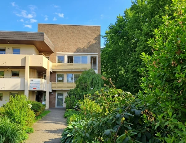 Auf dem Bild ist der Eingangsbereich eines mehrstöckigen Wohngebäudes mit Klinkerfassade und hellen Balkonen zu sehen. Davor führt ein gepflasterter Weg durch einen dicht bepflanzten, grünen Garten mit Sträuchern und Bäumen.