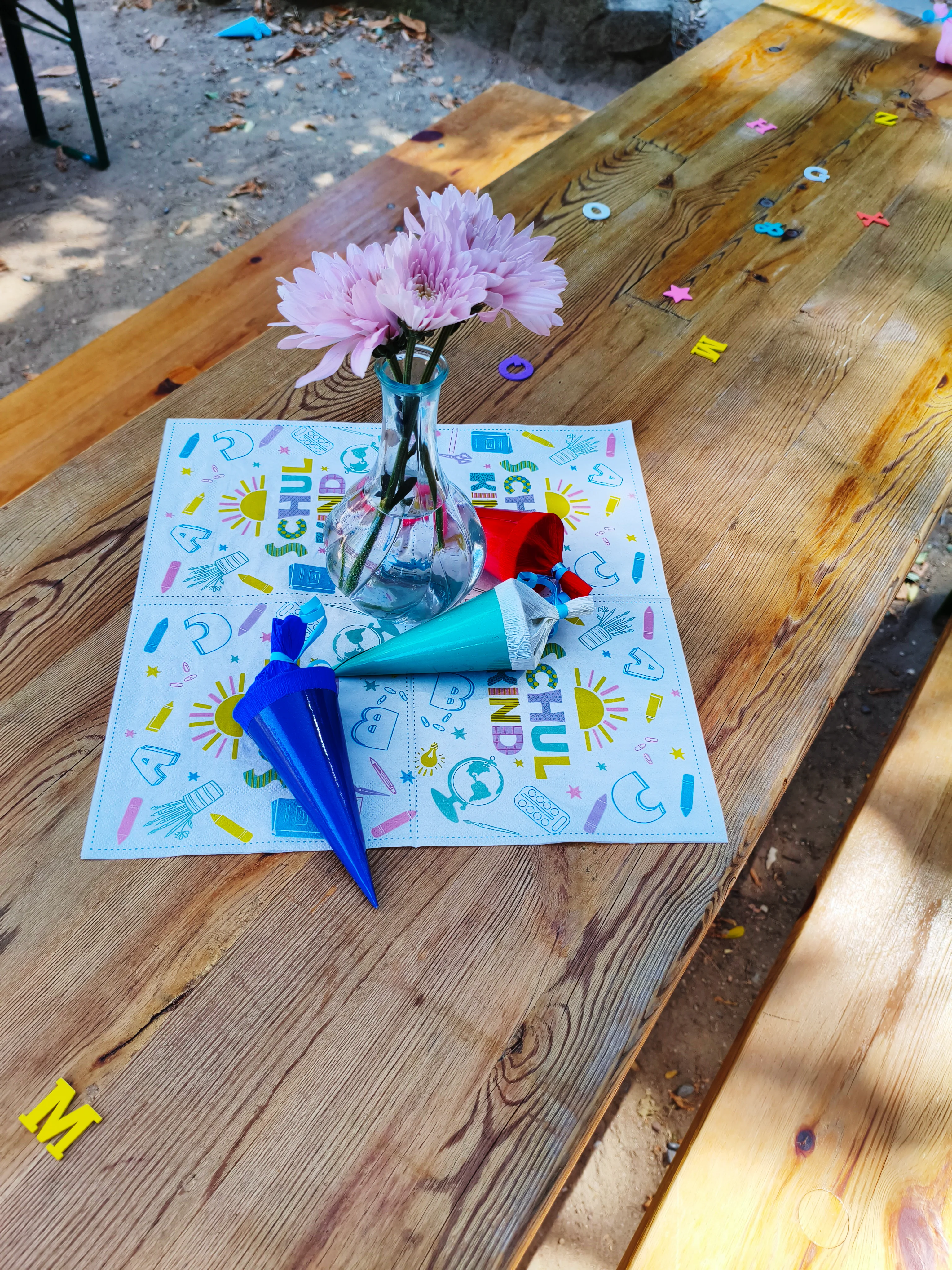 Auf dem Bild ist ein dekorierter Holztisch zu sehen, der offensichtlich für eine Einschulung oder ein Schulkind‑Fest vorbereitet ist. Motiv und Gegenstände Auf dem Tisch steht eine schmale Glasvase mit mehreren rosa Blüten, die zentral auf einer bunten Serviette platziert ist, auf der groß „Schulkind“ und typische Schulmotive wie Sonne, Globus und Stifte zu erkennen sind. Rund um die Vase liegen kleine, bunte Mini‑Schultüten sowie verstreute Kunststoff‑ oder Pappbuchstaben und Formen, die sich über den Tisch verteilen und für eine kindliche, verspielte Atmosphäre sorgen.