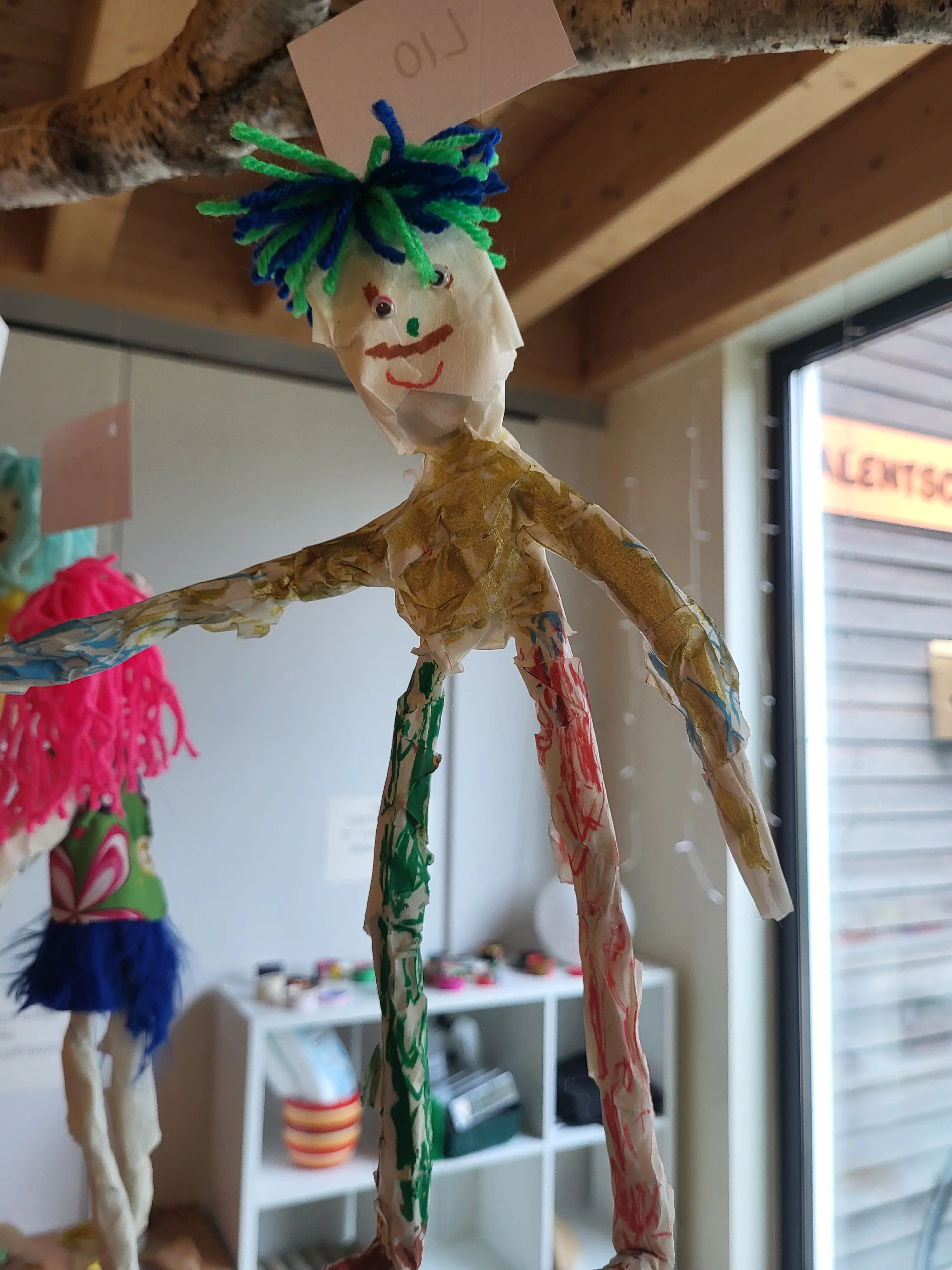 Auf dem Bild ist eine bunte, von Kindern gebastelte Figur aus Papier/Tape und Draht oder ähnlichem Material zu sehen, die an einem Ast von der Decke hängt. Die Figur hat Haare aus grüner und blauer Wolle, ein gemaltes Gesicht mit Mund und Nase sowie farbig bemalte Arme und Beine; oben hängt ein Schild mit dem Namen „Lio“.