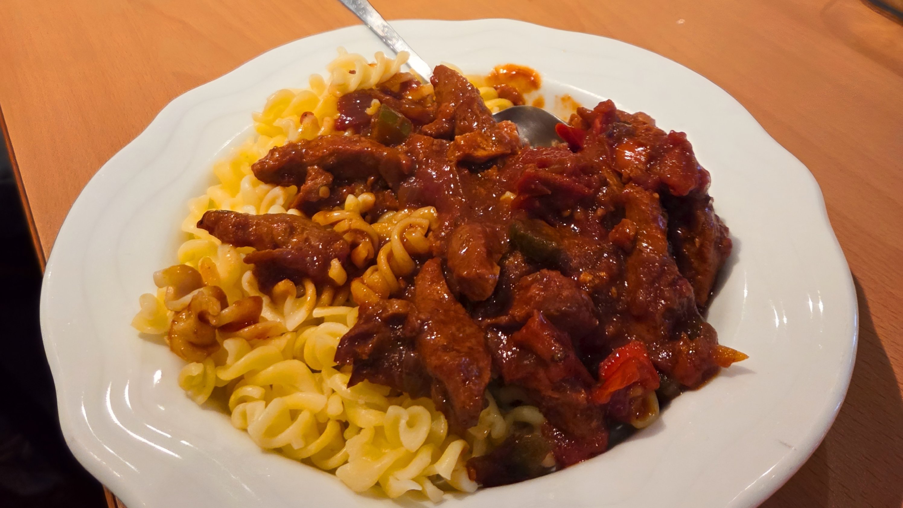 Auf dem Foto ist ein Teller mit spiraligen Nudeln zu sehen, darüber eine dunkle, tomatige Fleisch‑ oder Gulaschsoße mit Paprika- oder Gemüsestücken. Im Hintergrund sitzt eine kleine Runde am Tisch, in der Mitte steht ein großer Edelstahltopf sowie Gläser mit Wasser auf einem gedeckten Tisch mit roten Servietten.