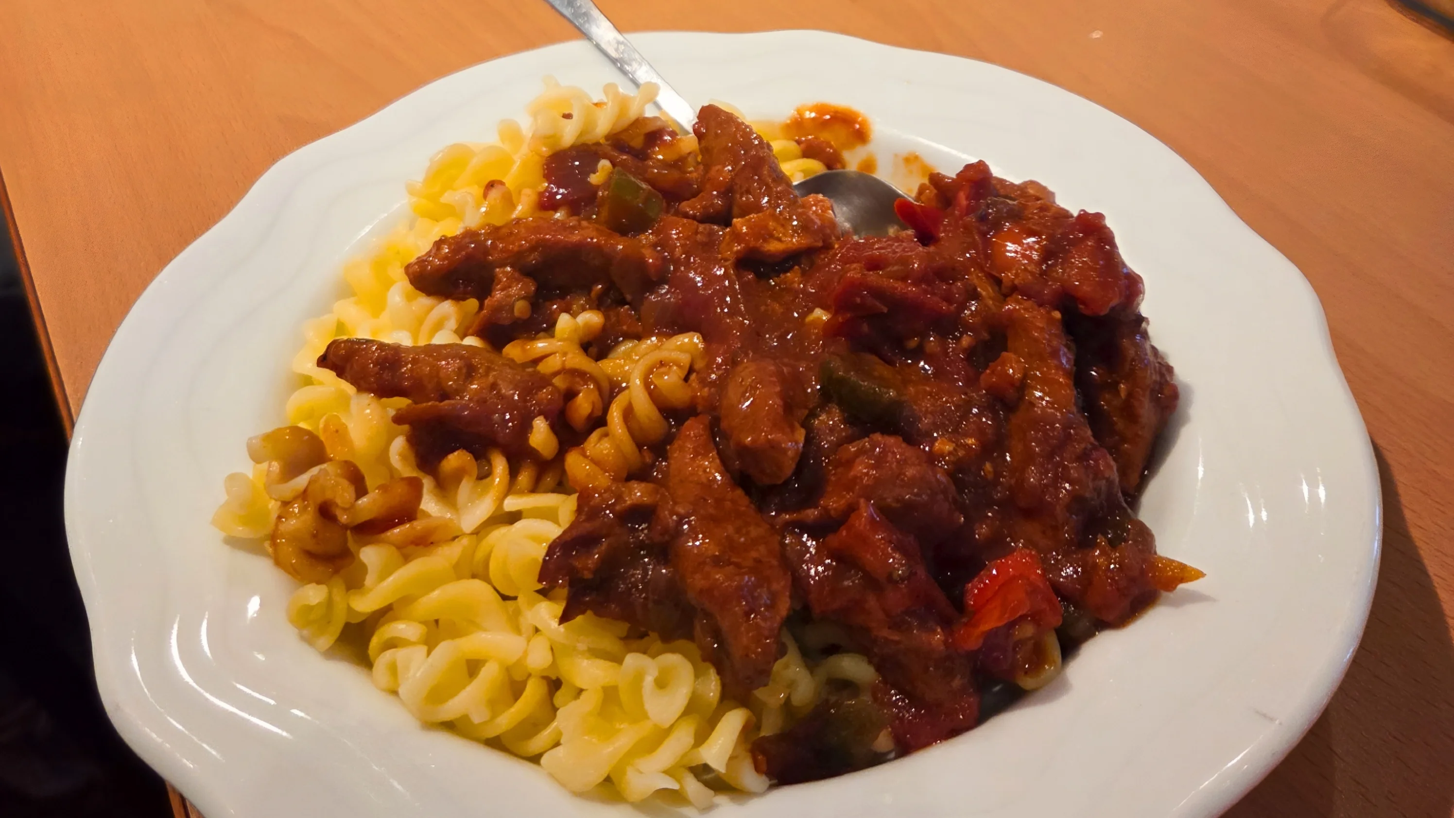 Auf dem Foto ist ein Teller mit spiraligen Nudeln zu sehen, darüber eine dunkle, tomatige Fleisch‑ oder Gulaschsoße mit Paprika- oder Gemüsestücken. Im Hintergrund sitzt eine kleine Runde am Tisch, in der Mitte steht ein großer Edelstahltopf sowie Gläser mit Wasser auf einem gedeckten Tisch mit roten Servietten.