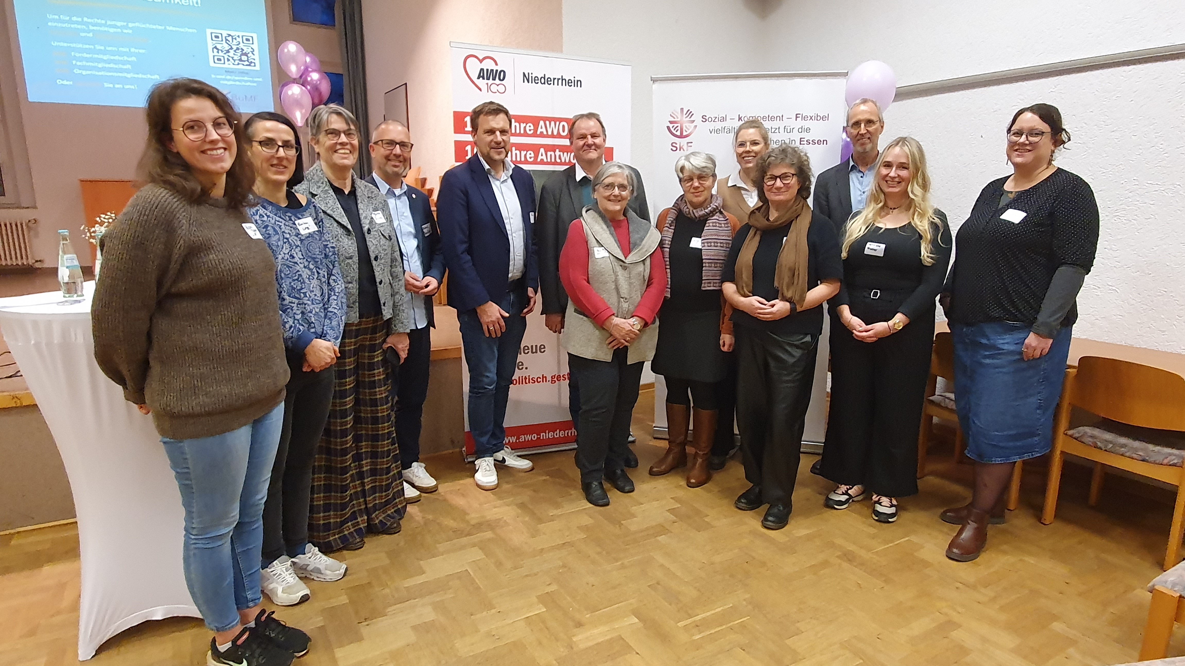Auf dem Bild ist eine Gruppe von 14 Erwachsenen zu sehen, die in einem Innenraum zusammenstehen. Es sieht nach einer Veranstaltung oder einem Treffen aus, möglicherweise im Rahmen der AWO Niederhein und der SKF (Sozialdienst katholischer Frauen). Personen: Die Gruppe besteht aus etwa sieben Frauen und sieben Männern, die in festlicher, aber nicht übermäßig formeller Kleidung posieren. Sie stehen in zwei lockeren Reihen beieinander. Hintergrund: Im Hintergrund sind mehrere Banner und Plakate zu sehen, die auf die Organisationen hinweisen: Ein Logo mit der Aufschrift "AWO 100 Niederhein 100 Jahre Antwort auf die soziale Frage" ist erkennbar. Ein weiteres Plakat trägt die Aufschrift "Sozial - Kompetent - Flexibel" und "SKF" sowie den Ort "Essen". Es sind auch einige lilafarbene und graue Luftballons im Raum verteilt. Vordergrund: Links im Bild steht ein mit einem weißen Tuch bedeckter Tisch. Der Boden ist ein heller Holzboden.