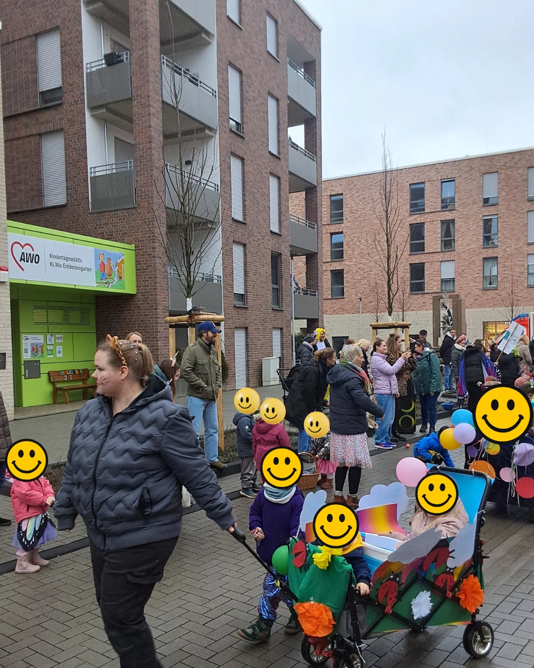 Auf dem Bild ist eine Karnevalsfeier oder ein Karnevalsumzug in einer Wohnsiedlung zu sehen. Im Vordergrund und in der Mitte des Bildes befinden sich mehrere Erwachsene und Kinder (deren Gesichter mit gelben Smiley-Emojis verdeckt wurden).