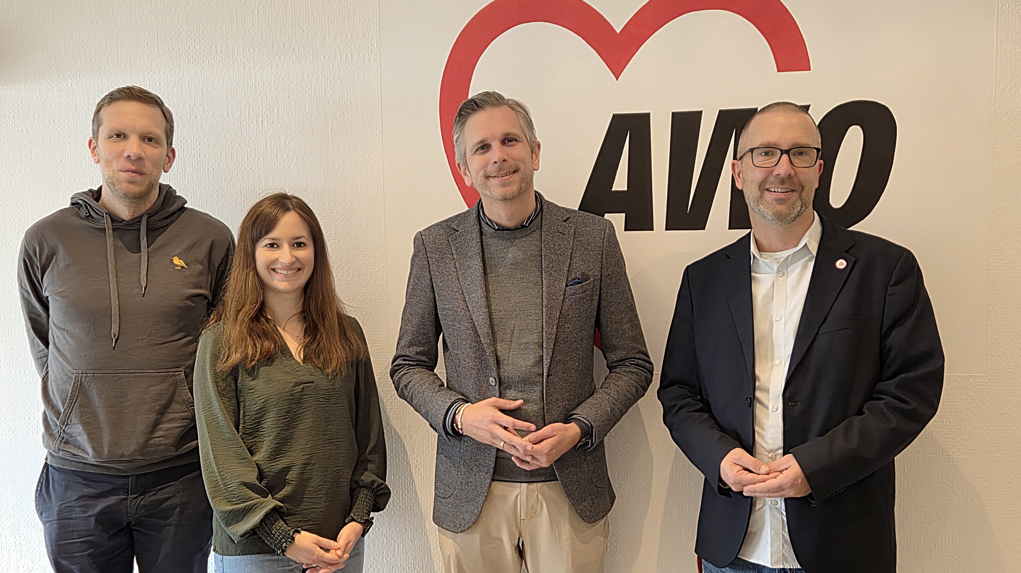 Auf dem Foto sind vier Personen vor einer weißen Wand mit einem großen Logo zu sehen. Drei der Personen sind Männer, und eine ist eine Frau. Sie stehen nebeneinander und lächeln. Hinter ihnen ist ein Logo zu sehen, das ein großes rotes Herz und die Buchstaben AWO enthält.