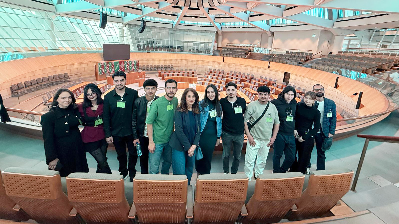 Auf dem Foto ist eine Gruppe von 12 Personen zu sehen, die in einem großen, kreisförmigen Raum mit vielen Sitzplätzen stehen. Der Raum hat eine hohe Decke mit einem auffälligen, sternförmigen Muster. Die Personen sind in legerer Kleidung gekleidet und scheinen ein informelles Treffen oder eine Führung zu haben. Die Sitzplätze sind in konzentrischen Kreisen angeordnet und es gibt eine kleine Bühne oder Plattform in der Mitte des Raumes. Die allgemeine Atmosphäre ist entspannt und freundlich.