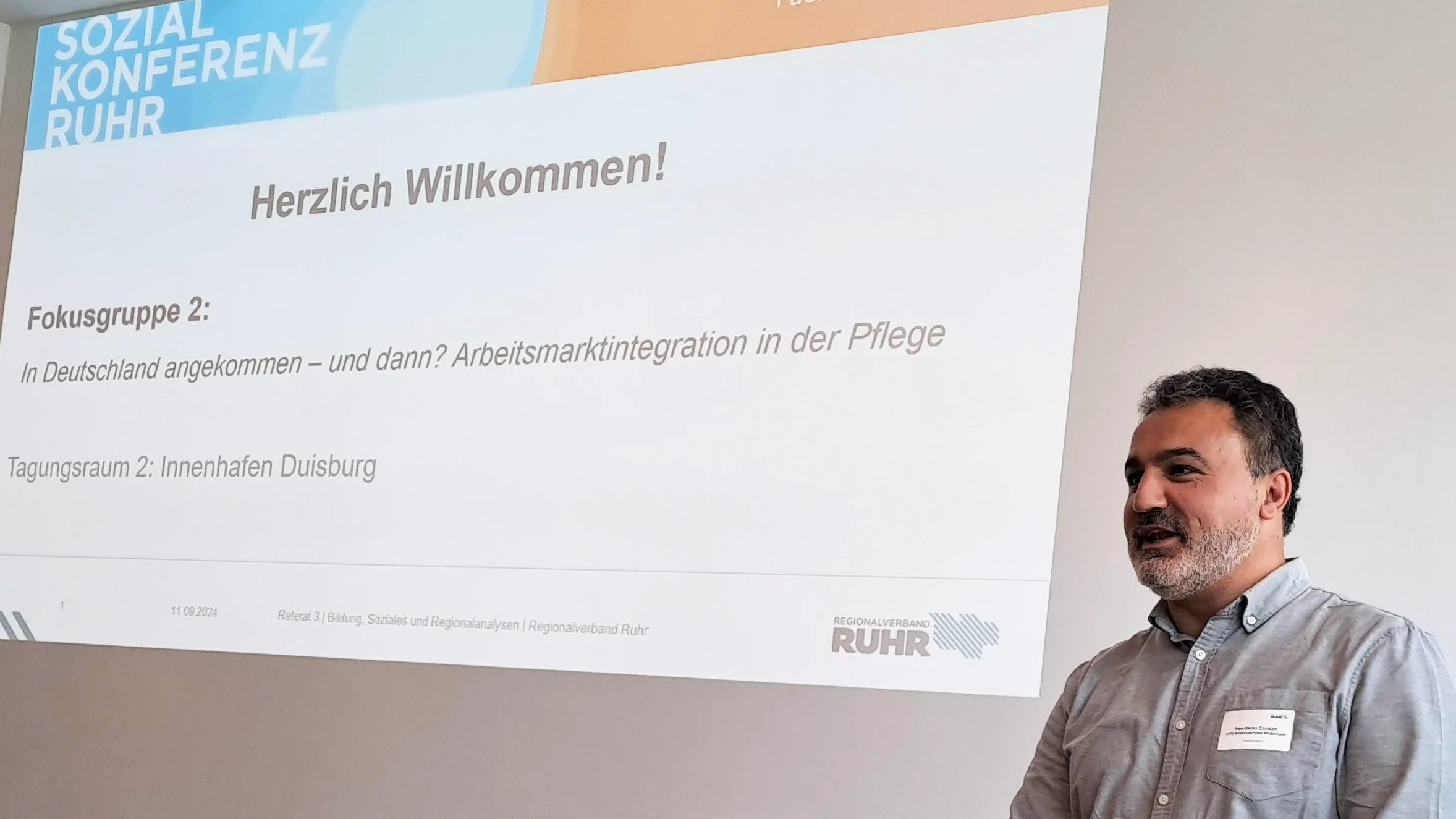 Das Foto zeigt eine Person, die vor einer Präsentationsleinwand steht. Auf der Leinwand steht “Herzlich Willkommen!” und es gibt Informationen zur “Sozialkonferenz RUHR, die sich mit den Themen Zukunft der Pflege im Ruhrgebiet und der Integration von Fachkräften beschäftigt. Die Person steht hinter einem Laptop und steuert vermutlich die Präsentation.
