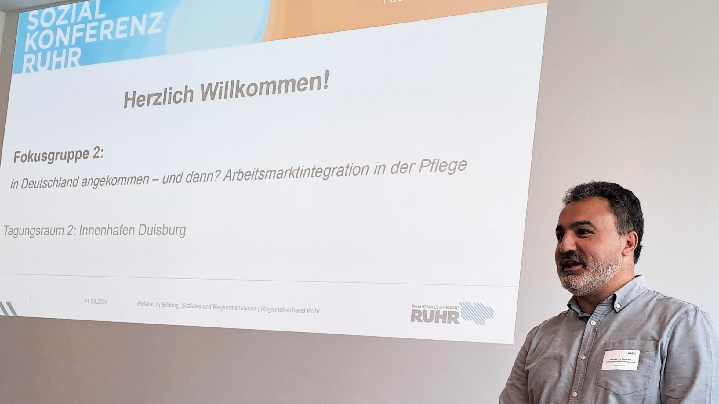 Das Foto zeigt eine Person, die vor einer Präsentationsleinwand steht. Auf der Leinwand steht “Herzlich Willkommen!” und es gibt Informationen zur “Sozialkonferenz RUHR, die sich mit den Themen Zukunft der Pflege im Ruhrgebiet und der Integration von Fachkräften beschäftigt. Die Person steht hinter einem Laptop und steuert vermutlich die Präsentation.