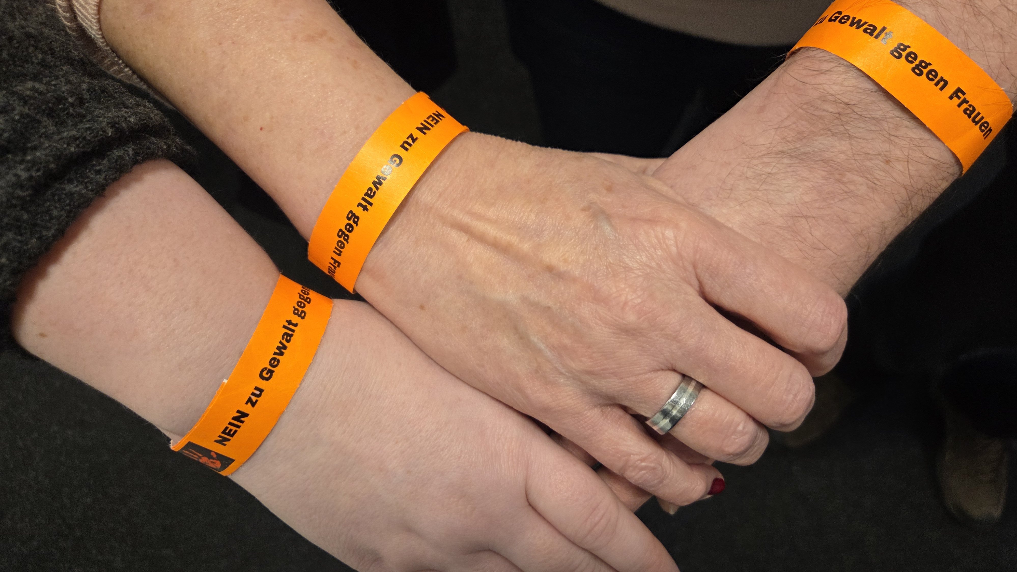 Auf dem Bild sind die Hände von drei Personen zu sehen, die ineinandergelegt sind. Drei Arme tragen orangefarbene Armbänder mit einer Aufschrift. Die Aufschrift, die man lesen kann, lautet: "NEIN zu Gewalt gegen Frauen". Die Hand in der Mitte trägt einen silbernen Ring am Ringfinger. Die Kleidung der Personen ist im Hintergrund angedeutet, unter anderem ein dunkler Ärmel und ein heller Oberteil.