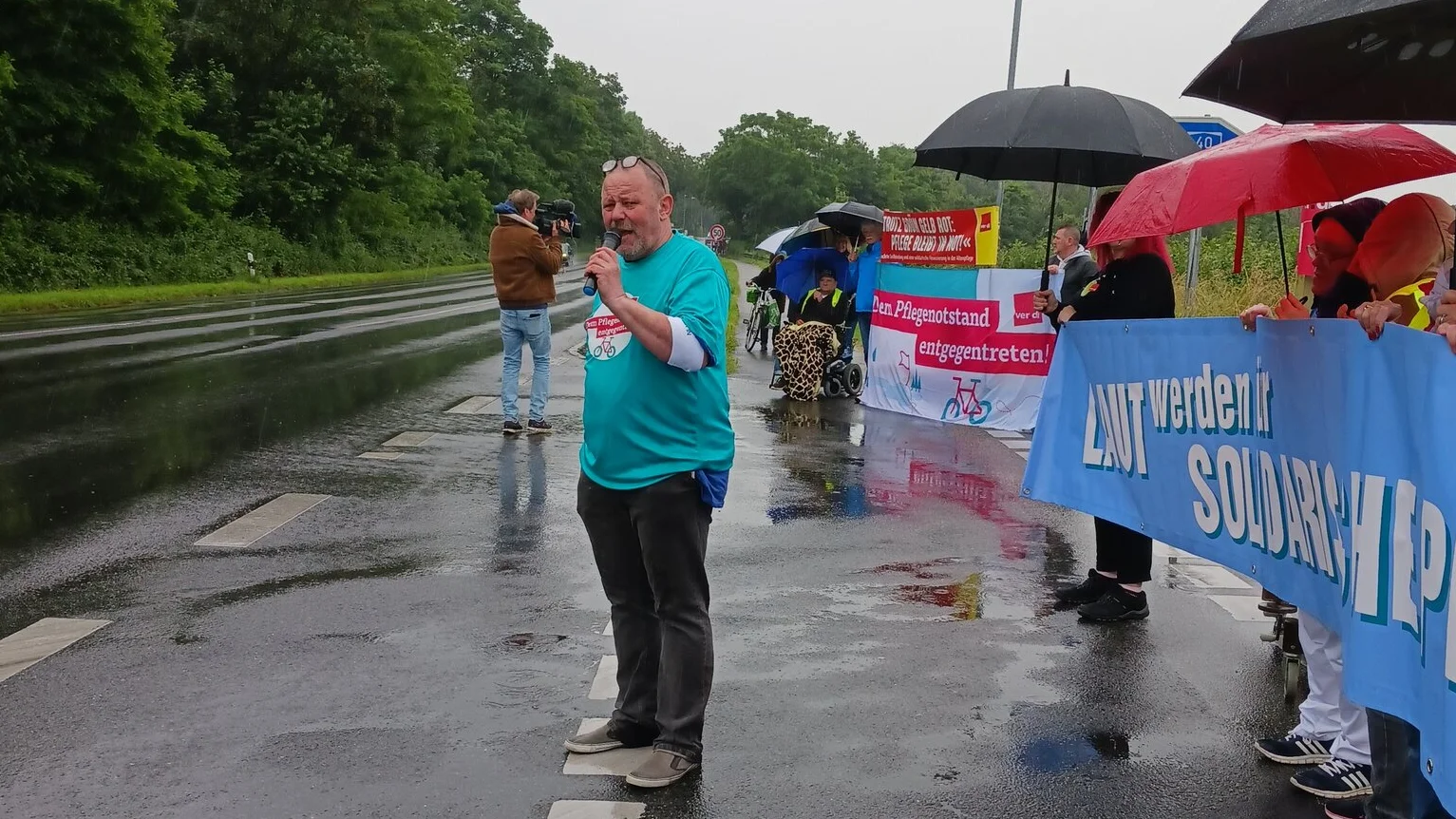 Auf dem Foto sind mehrere Personen zu sehen, die auf einer Straße stehen. Einige halten Regenschirme und Banner. Die Banner scheinen Text und möglicherweise Slogans zu enthalten, aber der genaue Inhalt ist aus dieser Perspektive nicht klar erkennbar. Die Straße ist nass, was auf kürzlichen Regen hindeutet, und es gibt Bäume auf beiden Seiten der Straße. Diese Szene weist auf eine Demonstration oder einen Protest auf der Straße hin.
