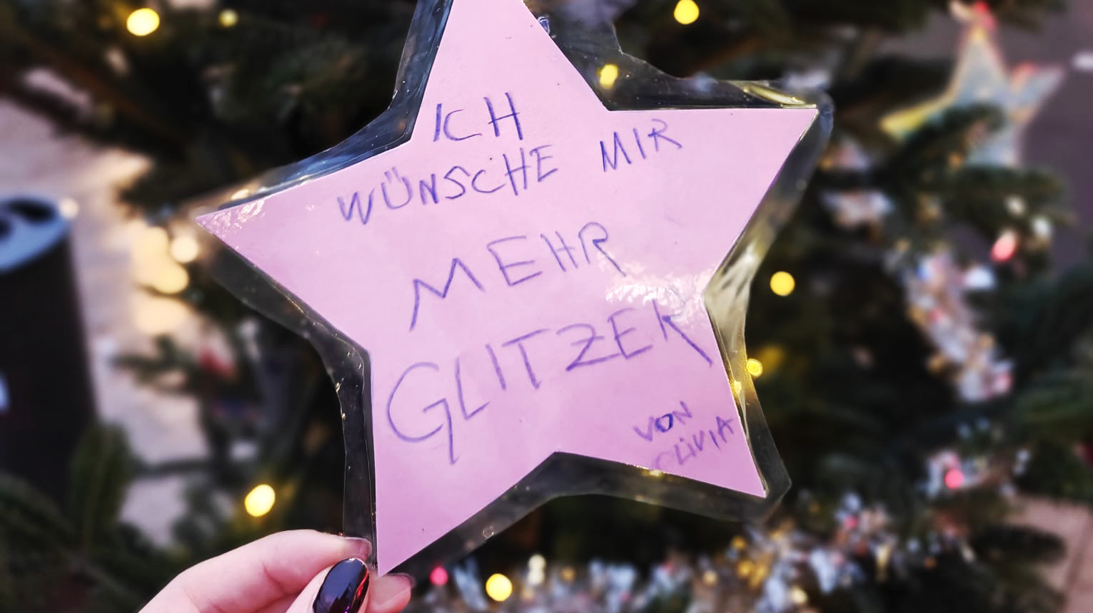 Auf dem Bild ist ein Detail eines Weihnachtsbaumes im Freien zu sehen, der mit Lichtern geschmückt ist. Im Vordergrund hängt ein großer, pinker Stern aus Pappe oder Papier, der laminiert zu sein scheint, als Christbaumschmuck an einem Ast. Der Stern ist mit einer Nachricht in lila/blauer Schrift beschriftet, die lautet: ICH WÜNSCHE MIR MEHR GLITZER von Livia Eine Hand mit dunkel lackierten Fingernägeln hält den Stern leicht von unten. Zusammenfassend zeigt das Bild einen Wunschstern, der an einem Tannenbaum hängt und von einem Kind namens Livia mehr Glitzer fordert.
