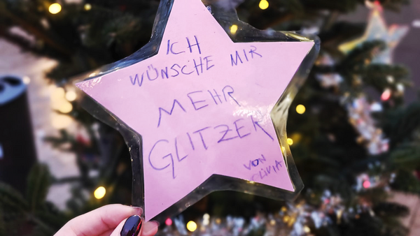 Auf dem Bild ist ein Detail eines Weihnachtsbaumes im Freien zu sehen, der mit Lichtern geschmückt ist. Im Vordergrund hängt ein großer, pinker Stern aus Pappe oder Papier, der laminiert zu sein scheint, als Christbaumschmuck an einem Ast. Der Stern ist mit einer Nachricht in lila/blauer Schrift beschriftet, die lautet: ICH WÜNSCHE MIR MEHR GLITZER von Livia Eine Hand mit dunkel lackierten Fingernägeln hält den Stern leicht von unten. Zusammenfassend zeigt das Bild einen Wunschstern, der an einem Tannenbaum hängt und von einem Kind namens Livia mehr Glitzer fordert.
