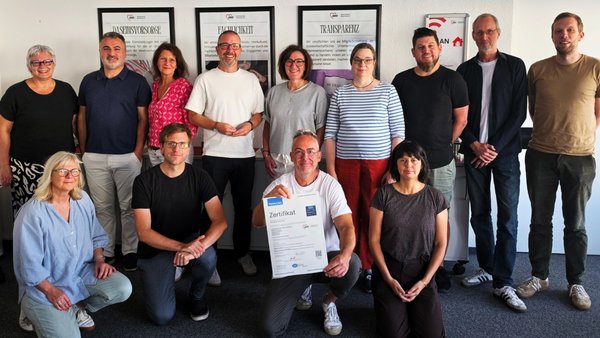 Zu sehen ist eine Gruppenaufnahme mit Mitarbeitenden beziehungsweise Abteilungsleitungen des AWO Bezirksverbands Niederrhein sowie dem Vorstand. Im Vordergrund hält ein Teilnehmer das neue TÜV-Zertifikat in die Kamera; im Hintergrund sind gerahmte AWO-Leitbildplakate zu Themen wie Daseinsvorsorge, Fachlichkeit und Transparenz angebracht.
