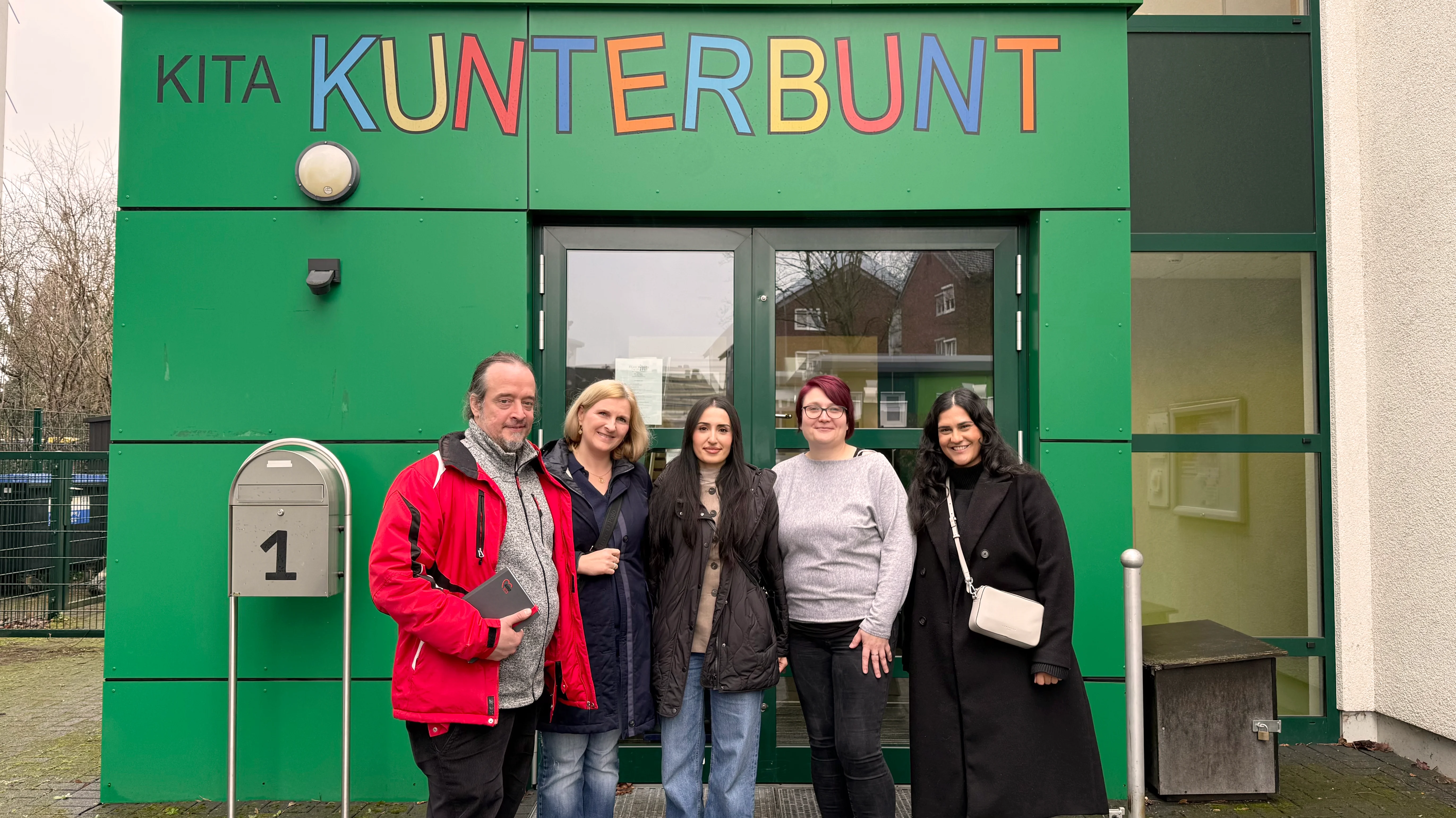 Auf dem Foto steht eine Gruppe von fünf Erwachsenen vor dem Eingang einer Kindertagesstätte mit der Aufschrift „KITA KUNTERBUNT“. Die Personen stehen dicht beieinander, tragen Herbst‑ bzw. Winterkleidung und blicken zur Kamera.
