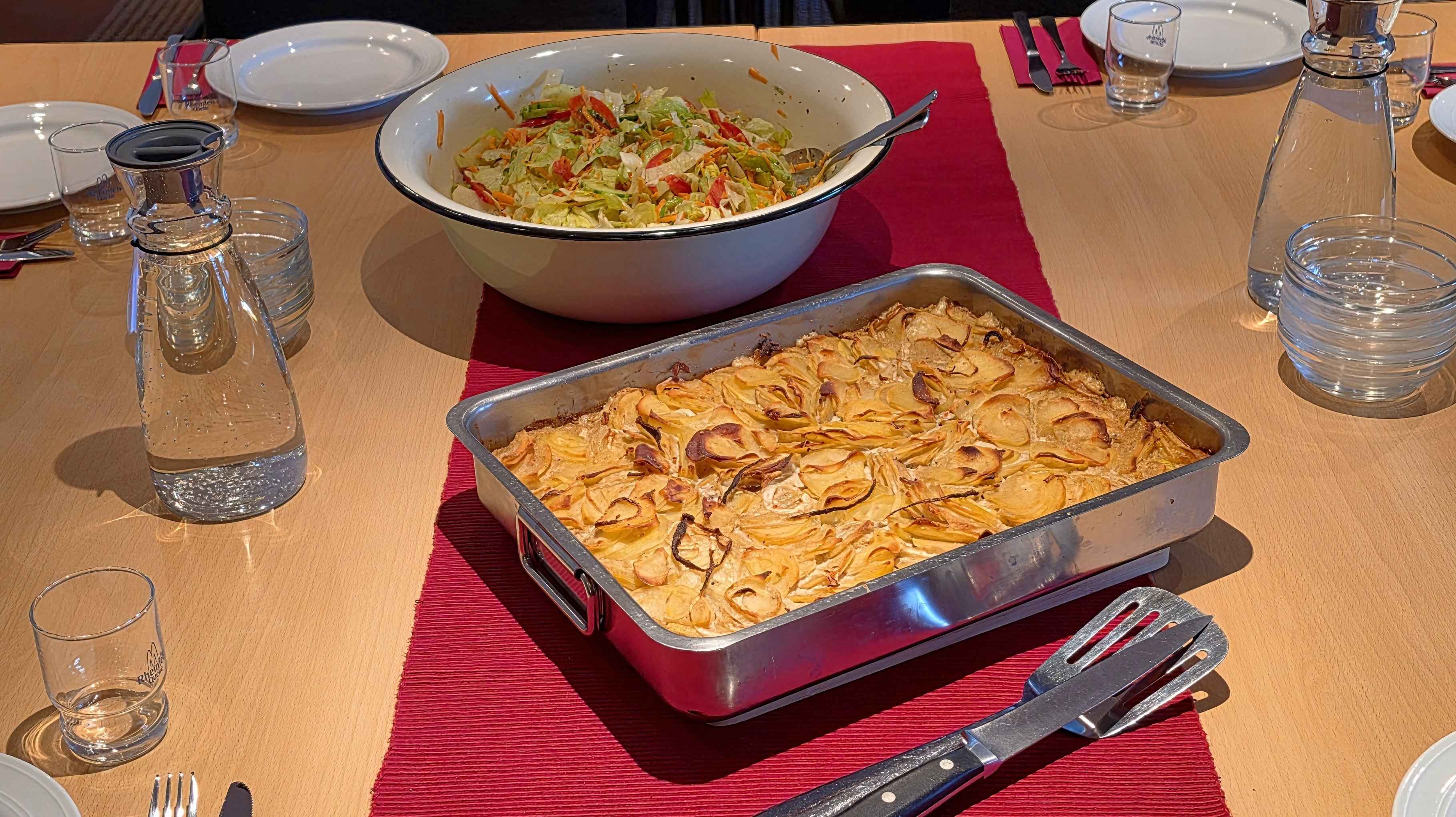 Auf dem Bild ist ein gedeckter Tisch in einem Besprechungs- oder Aufenthaltsraum zu sehen. In der Mitte stehen eine große Ofenform mit Kartoffelgratin und eine große Schüssel mit Salat, darum herum sind Teller, Besteck, Gläser und Wasserkaraffen für mehrere Personen angerichtet.