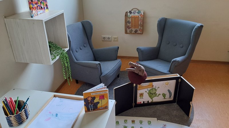 Auf dem Bild sieht man einen gemütlich eingerichteten Raum für Gespräche oder Spielangebote mit Kindern. Im Hintergrund stehen zwei gepolsterte, graublaue Sessel auf einem runden Teppich, dazwischen ein kleiner Tisch oder Hocker. Davor ist ein Kamishibai‑Erzähltheater mit einer Bildergeschichte zu Monstern oder Gefühlen aufgebaut, daneben liegen großformatige Bildkarten auf dem Boden. Im Vordergrund steht ein kleiner Tisch mit einem Kinderbild auf einem Holztablett, einem Becher mit bunten Stiften und einem kleinen aufgeschlagenen Heft. An der Wand hängen Regale mit einem Buch („Alle Zahlen“) und einer herunterhängenden Grünpflanze sowie weitere dekorative Elemente und kleine Pflanzen auf der Fensterbank.