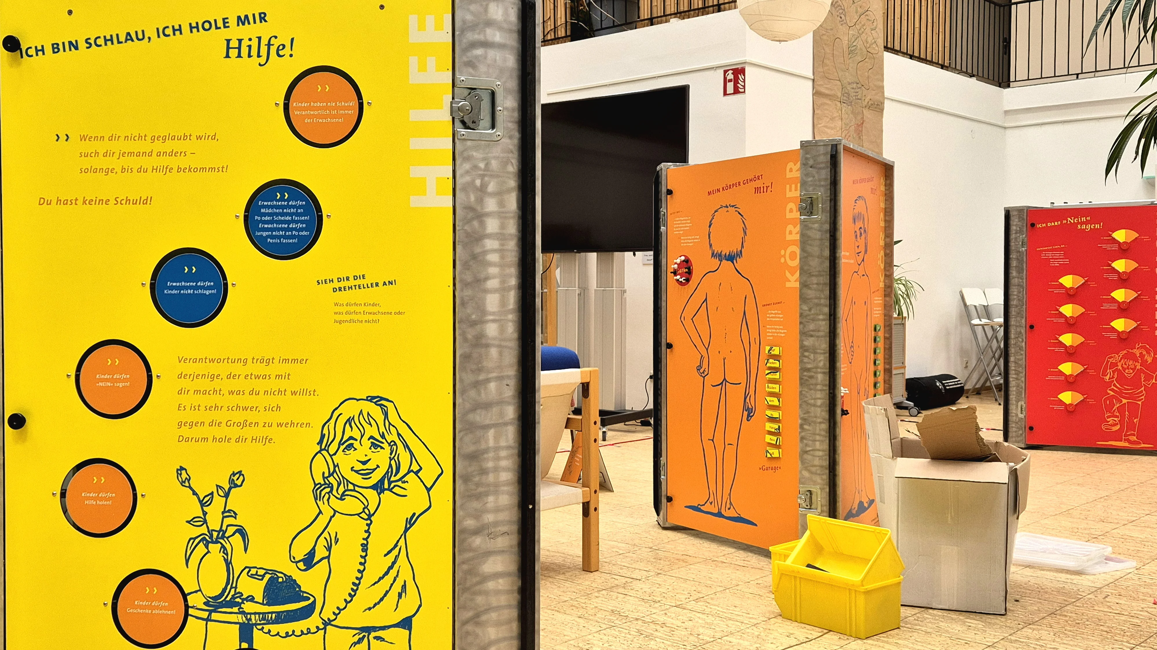 Das Foto zeigt eine helle Innenumgebung mit einem auffälligen gelben Informationsboard im Vordergrund links. Auf dem Board sind verschiedene Texte und Illustrationen zu sehen, darunter eine Zeichnung eines Mädchens mit Sprechblasen. Es gibt auch eine orangefarbene Figur mit Text, die in der Nähe steht. Im Hintergrund befindet sich ein weiteres Informationsboard in Rot.