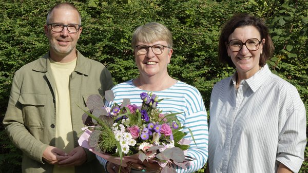Auf dem Foto sind drei lächelnde Erwachsene vor dichtem grünen Gebüsch zu sehen. Die Frau in der Mitte hält einen bunten Blumenstrauß mit rosa, weißen und violetten Blüten. Alle drei tragen Brillen; links steht ein Mann, rechts eine Frau.