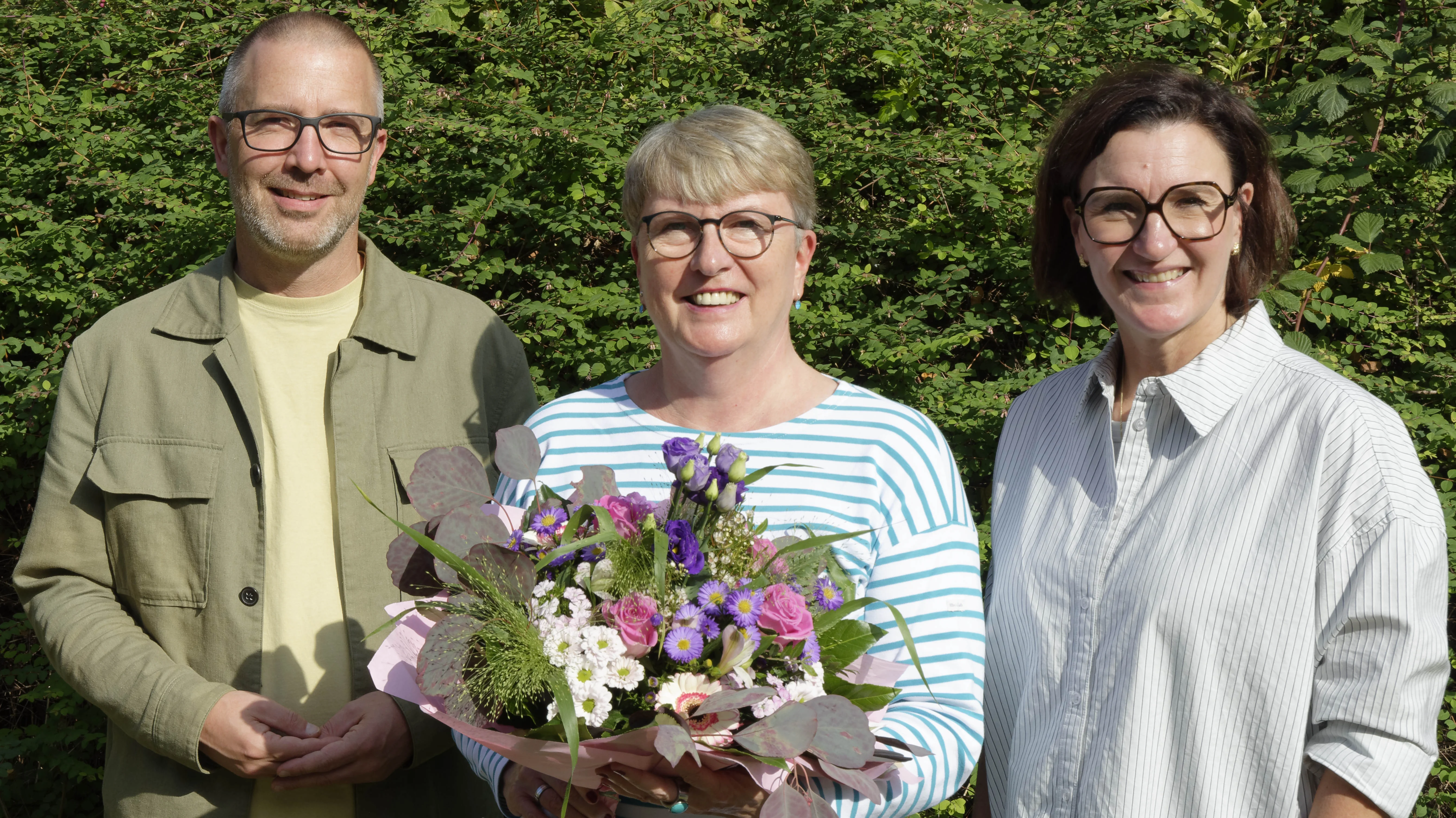 Auf dem Foto sind drei lächelnde Erwachsene vor dichtem grünen Gebüsch zu sehen. Die Frau in der Mitte hält einen bunten Blumenstrauß mit rosa, weißen und violetten Blüten. Alle drei tragen Brillen; links steht ein Mann, rechts eine Frau.