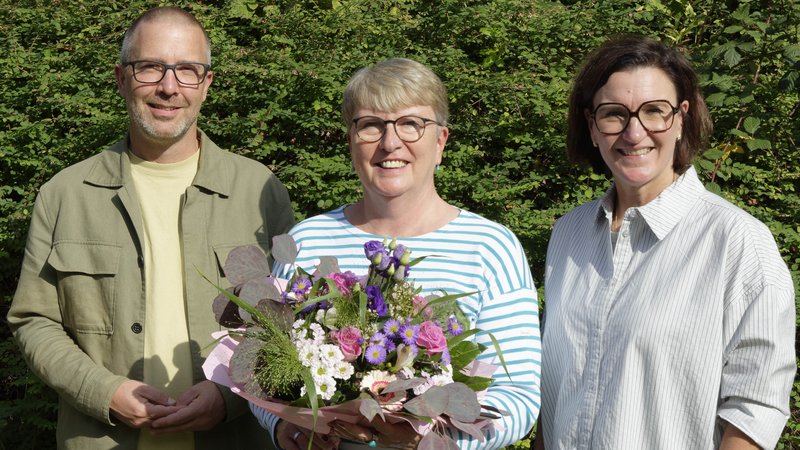 Auf dem Foto sind drei lächelnde Erwachsene vor dichtem grünen Gebüsch zu sehen. Die Frau in der Mitte hält einen bunten Blumenstrauß mit rosa, weißen und violetten Blüten. Alle drei tragen Brillen; links steht ein Mann, rechts eine Frau.