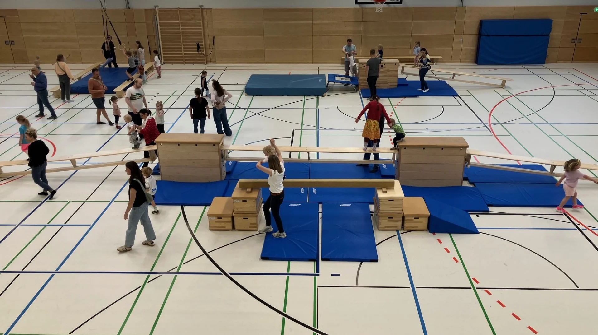 Auf dem Bild ist eine große Turnhalle zu sehen, in der viele Kinder gemeinsam mit Erwachsenen an einem Bewegungsparcours teilnehmen. Man erkennt verschiedene Turngeräte wie Kästen, Bänke und Matten, über die die Kinder balancieren, klettern und laufen, während die Erwachsenen daneben stehen, helfen oder zuschauen.