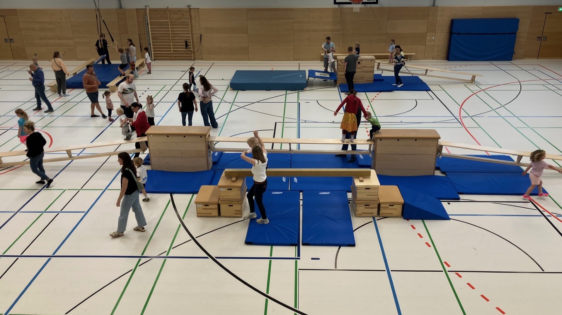 Auf dem Bild ist eine große Turnhalle zu sehen, in der viele Kinder gemeinsam mit Erwachsenen an einem Bewegungsparcours teilnehmen. Man erkennt verschiedene Turngeräte wie Kästen, Bänke und Matten, über die die Kinder balancieren, klettern und laufen, während die Erwachsenen daneben stehen, helfen oder zuschauen.