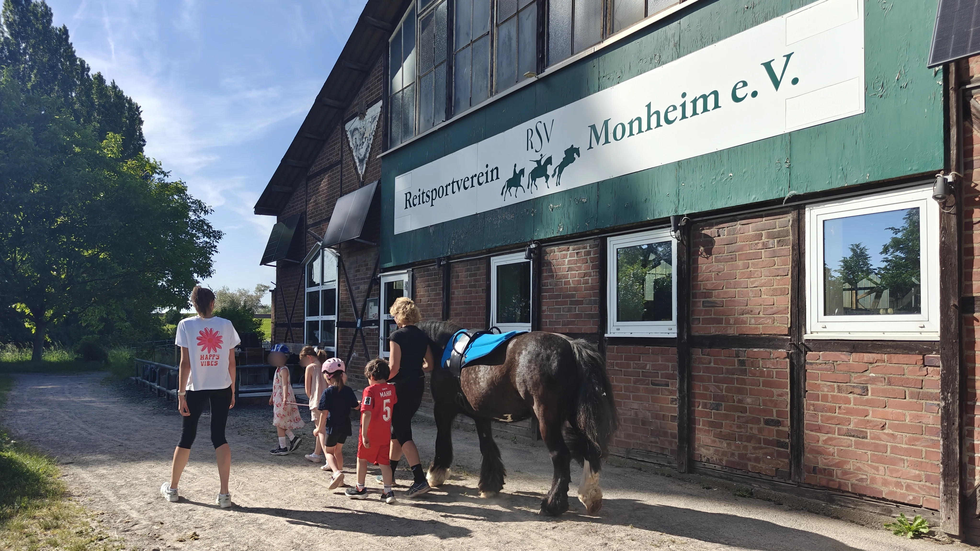 Mehrere Kinder gehen gemeinsam mit zwei Erwachsenen neben dem gesattelten Pony her. Im Hintergrund sieht man die Fassade eines Reitstalls mit dem Schriftzug „Reitsportverein Monheim e. V.“. Die Gruppe bewegt sich einen Weg entlang, es wirkt wie ein angeleitetes Angebot oder Ausflug auf dem Reiterhof.