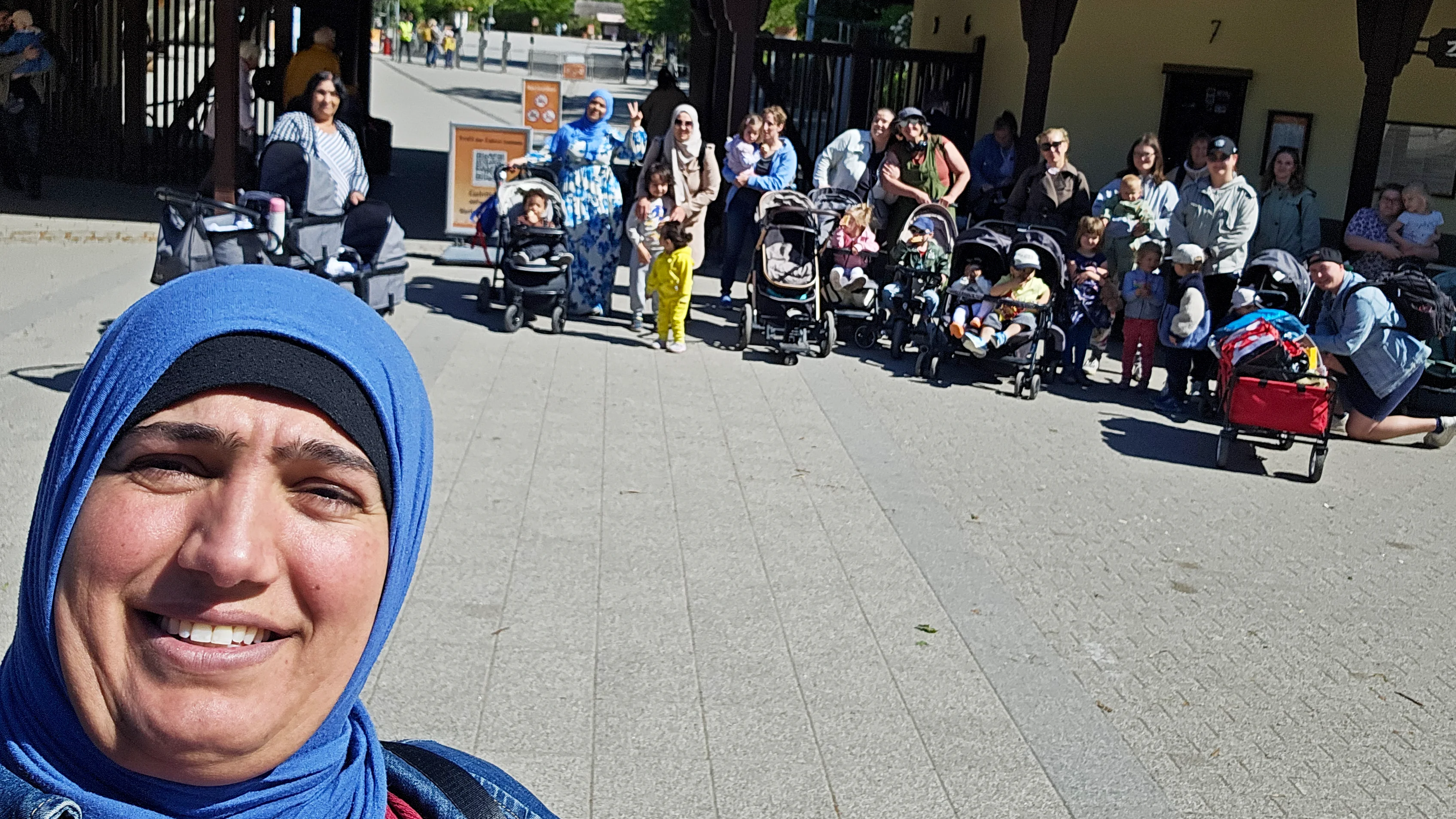 Auf dem Bild ist eine größere Gruppe Erwachsener mit vielen kleinen Kindern und mehreren Kinderwagen zu sehen, die vor einem Eingangsbereich mit überdachtem Tor steht. Im Vordergrund ist eine Person mit blauem Kopftuch im Selfie-Stil angeschnitten, im Hintergrund wirkt es wie der Vorplatz eines Zoos oder Parks.
