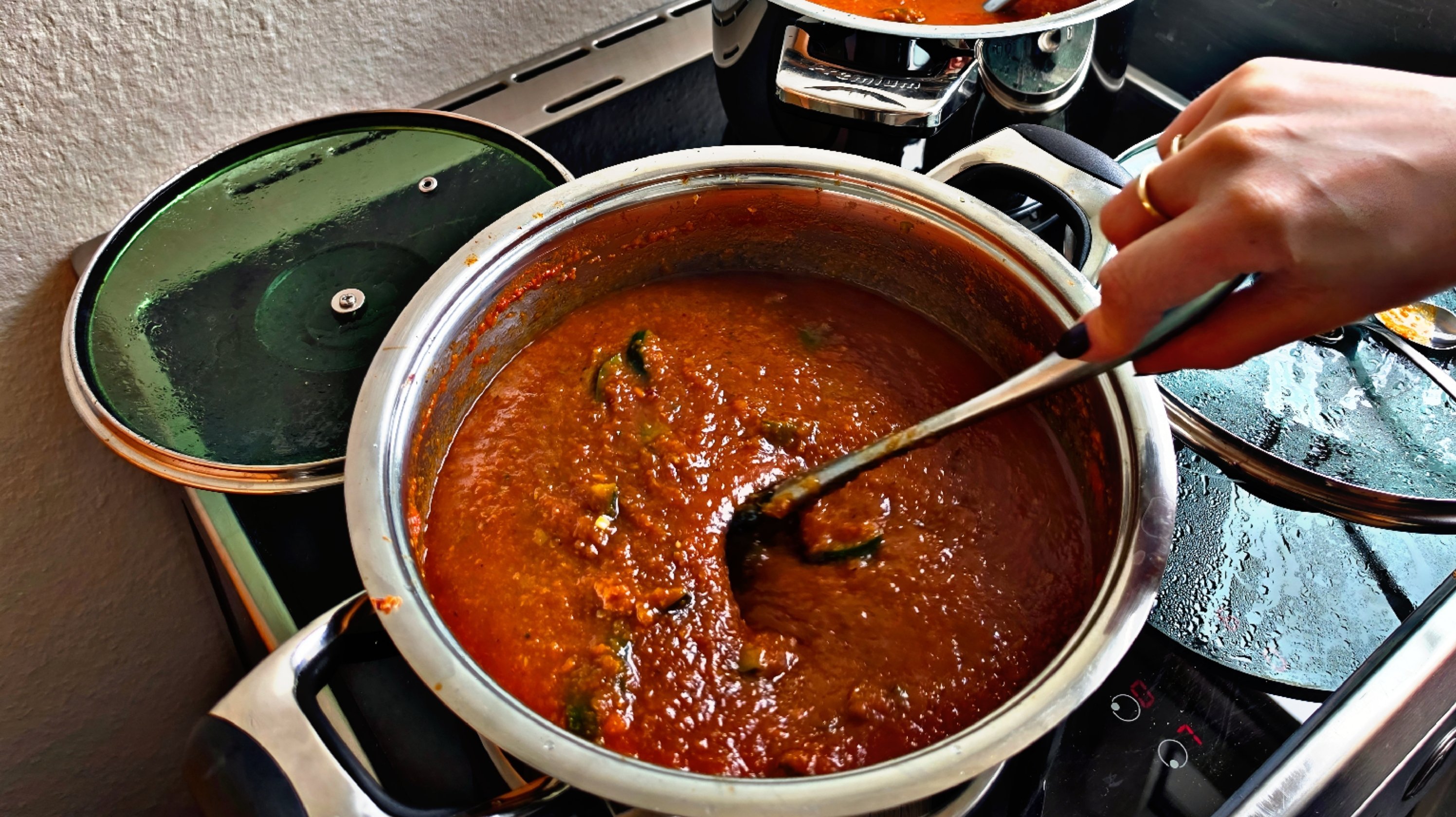 Auf dem Bild sieht man einen Herd mit zwei Töpfen, in denen eine rote, dicke Sauce oder Suppe kocht (vermutlich Tomatensauce oder Bolognese). In dem vorderen Topf rührt eine Hand mit einem Löffel um, daneben liegt ein grüner Topfdeckel.