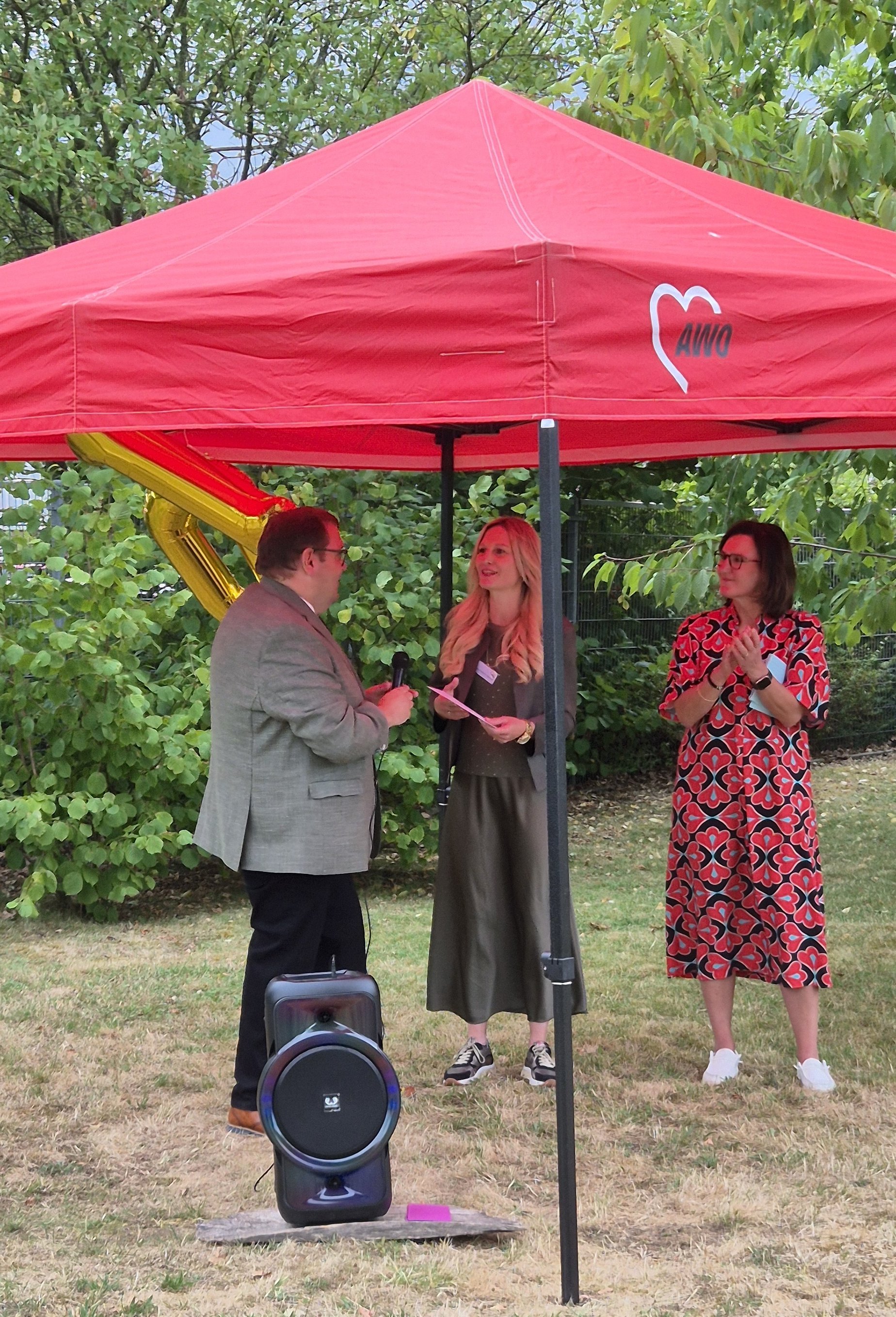 Drei Personen stehen unter einem roten Pavillon. Eine Person spricht in ein Mikrofon, während zwei weitere zuhören. Im Hintergrund sind grüne Büsche und Bäume sichtbar