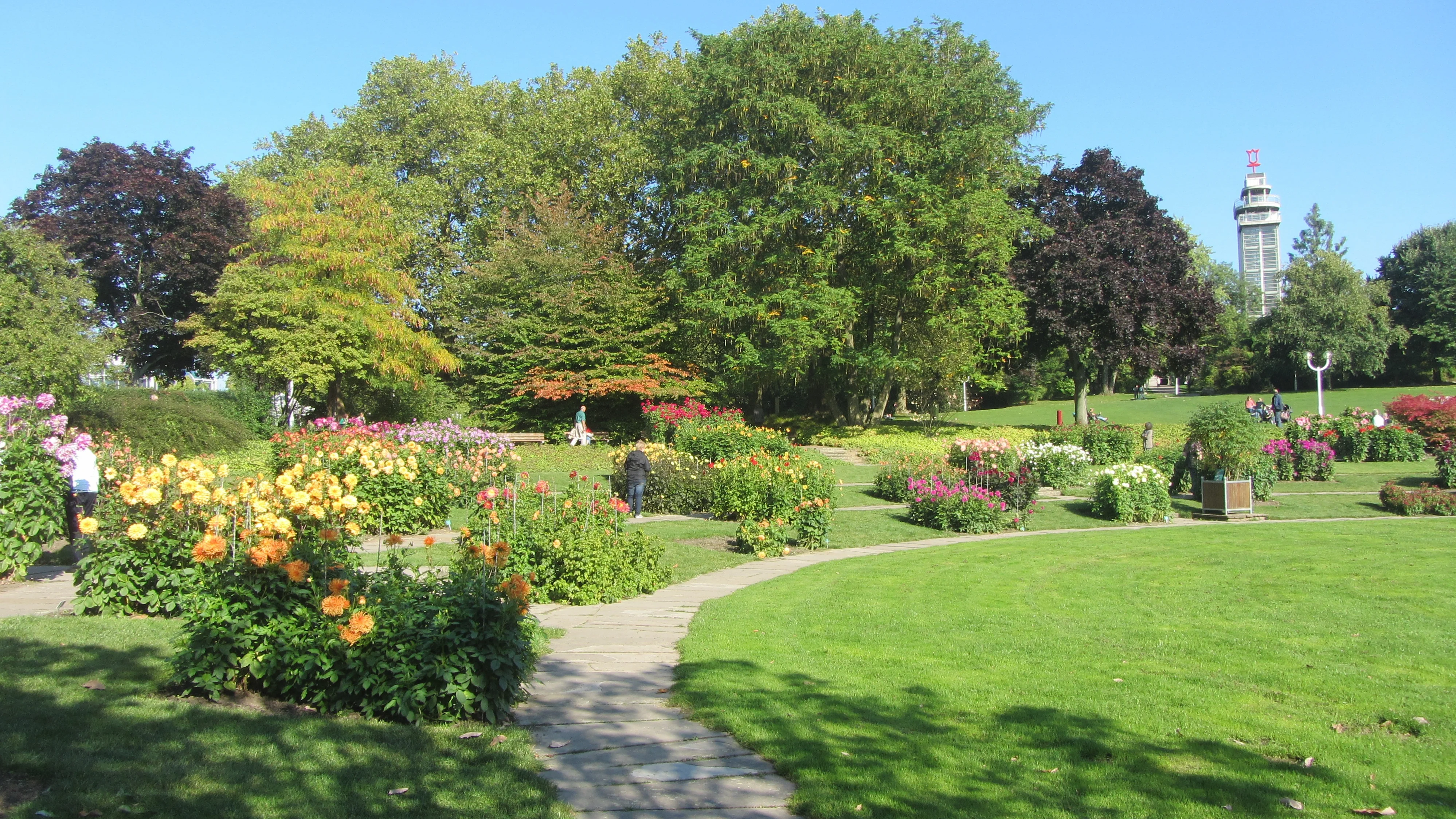 Auf dem Bild ist ein weitläufiger, gepflegter Park mit Blumenbeeten und großen Bäumen zu sehen. Einige Personen spazieren oder stehen zwischen den Beeten auf einem geschwungenen Weg, und im Hintergrund ragt ein markanter Aussichtsturm oder Gebäude mit einem Zeichen oben darauf über die Baumkronen empor.