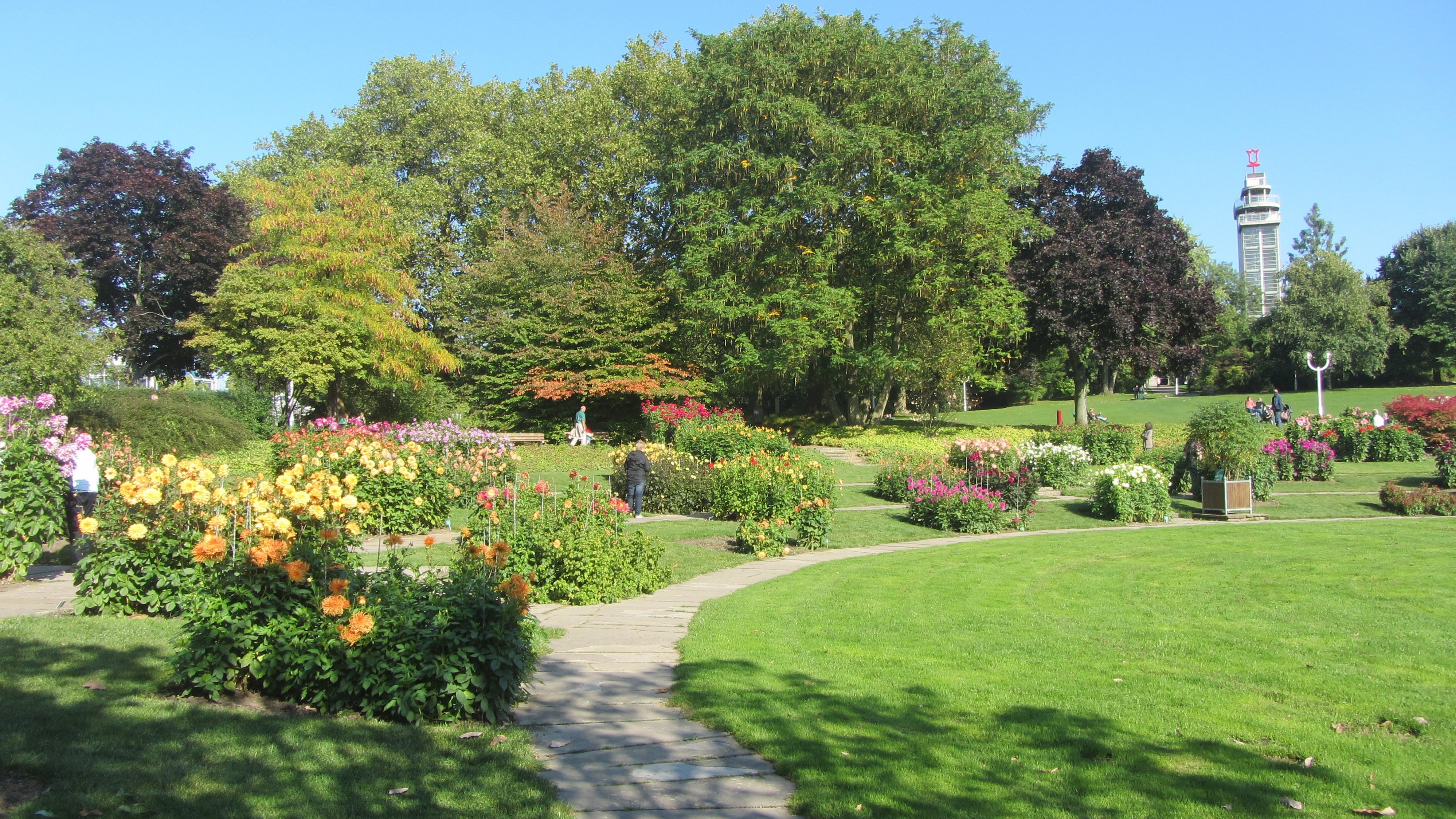 Auf dem Bild ist ein weitläufiger, gepflegter Park mit Blumenbeeten und großen Bäumen zu sehen. Einige Personen spazieren oder stehen zwischen den Beeten auf einem geschwungenen Weg, und im Hintergrund ragt ein markanter Aussichtsturm oder Gebäude mit einem Zeichen oben darauf über die Baumkronen empor.