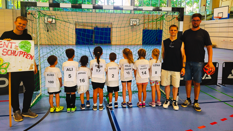 Auf dem Foto sieht man eine Gruppe von Kindern und drei erwachsene Betreuer in einer Sporthalle vor einem Handballtor. Die Kinder stehen mit dem Rücken zur Kamera und tragen weiße Trikots mit großen Namens- und Nummernzetteln, etwa „ELIAS 100“, „ALI 5“ oder „LENA 8“. Links hält ein Betreuer ein bunt gestaltetes Schild mit der Aufschrift „Talentschuppen“ (der Anfang des Wortes ist angeschnitten), rechts hält ein anderer Betreuer einen kleinen Ball in der Hand. Im Hintergrund ist ein Hallennetz, Basketballkörbe und große Fensterfronten mit Blick ins Grüne zu sehen.