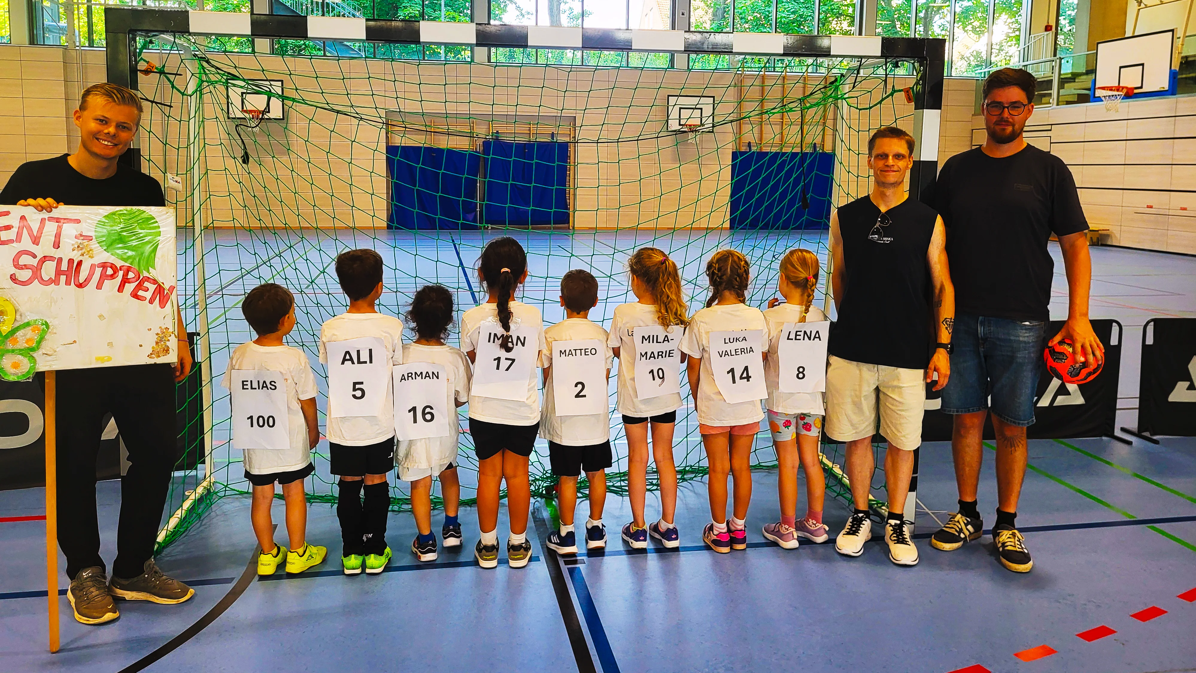 Auf dem Foto sieht man eine Gruppe von Kindern und drei erwachsene Betreuer in einer Sporthalle vor einem Handballtor. Die Kinder stehen mit dem Rücken zur Kamera und tragen weiße Trikots mit großen Namens- und Nummernzetteln, etwa „ELIAS 100“, „ALI 5“ oder „LENA 8“. Links hält ein Betreuer ein bunt gestaltetes Schild mit der Aufschrift „Talentschuppen“ (der Anfang des Wortes ist angeschnitten), rechts hält ein anderer Betreuer einen kleinen Ball in der Hand. Im Hintergrund ist ein Hallennetz, Basketballkörbe und große Fensterfronten mit Blick ins Grüne zu sehen.
