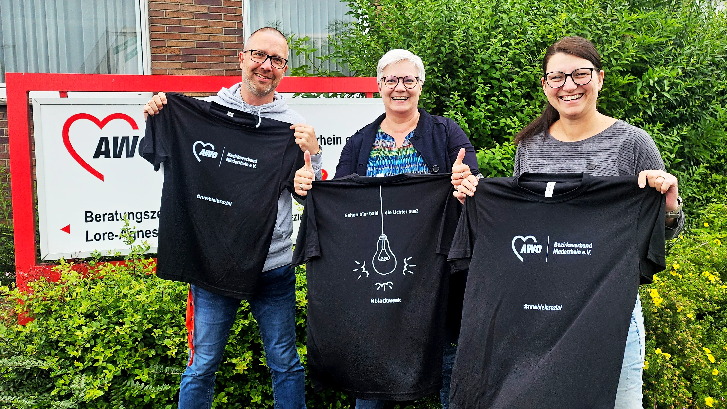 Das Foto zeigt drei Personen vor einem Gebäude mit einem roten und weißen Schild, auf dem ein AWO Logo zu sehen ist. Jede Person hält ein schwarzes T-Shirt mit weißem Text und Grafiken in der Hand. Auf den T-Shirts ist sowohl das Logo des AWO Bezirksverbands Niederrhein mit dem Hashtag nrwbleibsozial als auch eine Glühbirne mit dem Text „Gehen hier bald die Lichter aus?“ und dem Hashtag #blackweek zu sehen.