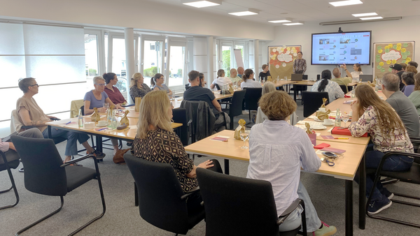 Das Foto zeigt einen Workshop in einem hellen, modernen Konferenzraum. Mehrere Personen sitzen an Tischen, die in Reihen aufgestellt sind, und schauen nach vorne zu einem Redner oder einer Rednerin, der oder die vor einer großen Leinwand steht. Auf der Leinwand ist eine Präsentation oder eine geteilte Bildschirminhalte zu sehen. Die Teilnehmenden haben Getränke und Schreibmaterial vor sich auf den Tischen liegen. Die Atmosphäre ist professionell und konzentriert.