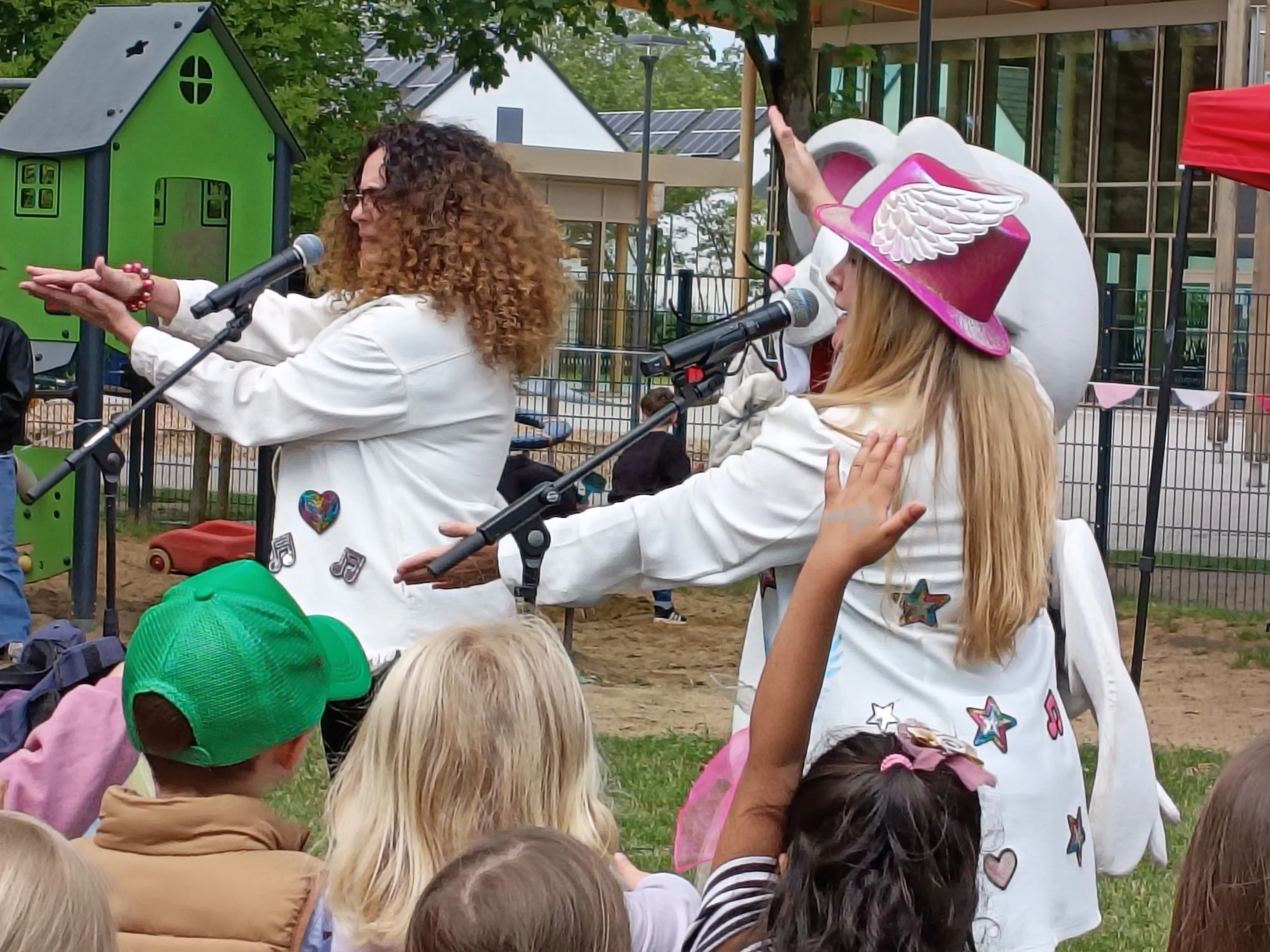 Auf dem Bild ist eine Live‑Kinderveranstaltung im Freien zu sehen. Personen und Aktion Im Vordergrund stehen zwei auftretende Frauen an Mikrofonen, beide in weißen Jacken mit bunten Symbolen (Herzen, Sterne, Noten). Die Frau rechts trägt einen pinken Hut mit weißen Flügeln; beide machen Gesten mit den Armen, die die Kinder nachahmen. Umgebung und Publikum Vor der Bühne sitzen bzw. stehen mehrere Kinder, die die Bewegungen mit erhobenen Armen mitmachen. Im Hintergrund erkennt man einen Spielplatz mit Kletterhaus sowie ein Gebäude mit großen Fenstern