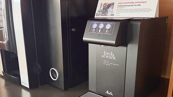 Auf dem Foto sind zwei Getränkeautomaten zu sehen. Einer davon ist ein schwarzer Automat mit der Aufschrift "fresh at work" und "COFFEE AND WATER AT WORK". Auf einem Schild darüber steht "Jetzt nachhaltig erfrischen! Trinkwasserspender für Alle". Auf dem Tresen vor dem Automaten steht ein Teller mit ein paar grünen und blauen Trauben.