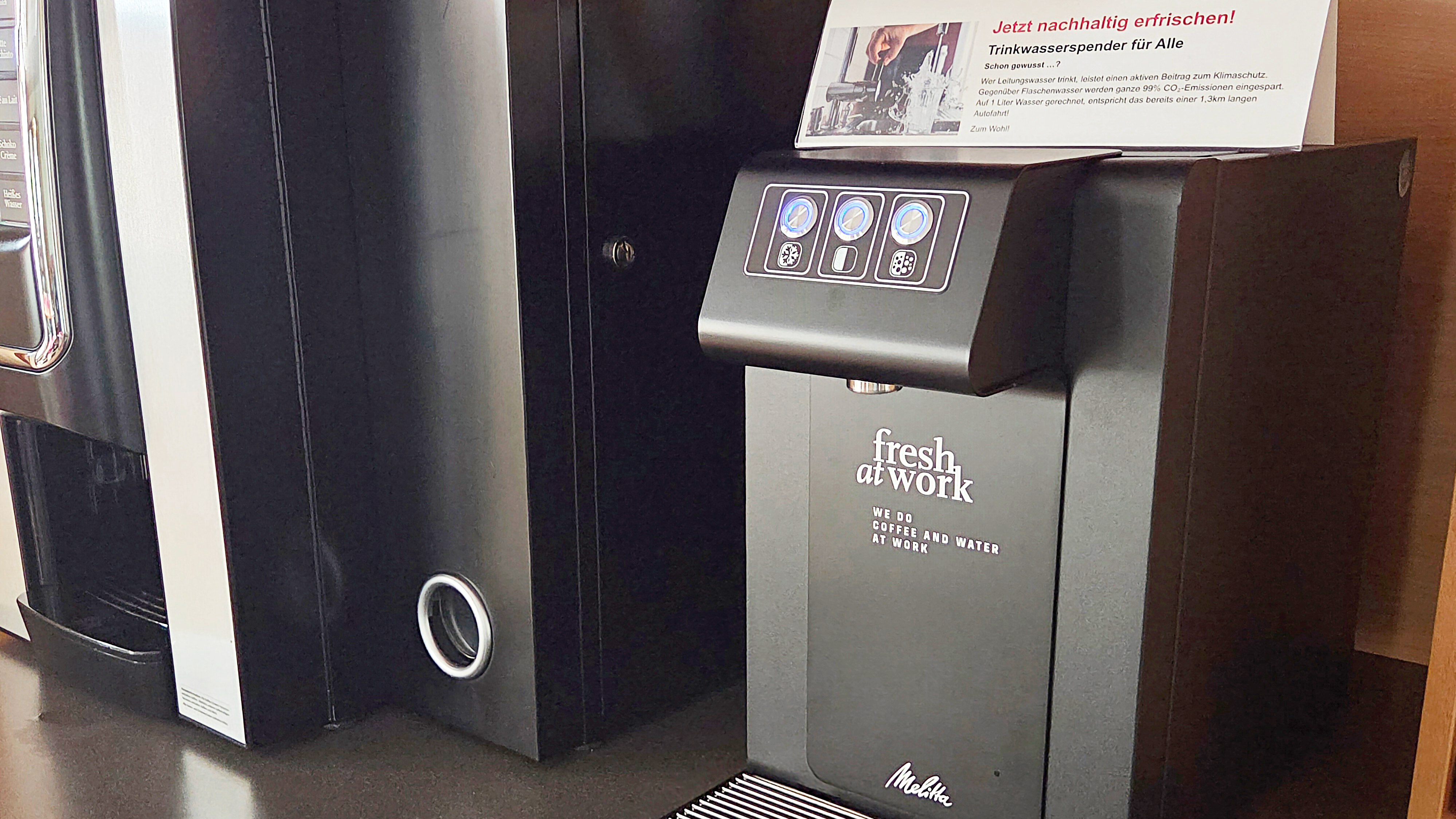 Auf dem Foto sind zwei Getränkeautomaten zu sehen. Einer davon ist ein schwarzer Automat mit der Aufschrift "fresh at work" und "COFFEE AND WATER AT WORK". Auf einem Schild darüber steht "Jetzt nachhaltig erfrischen! Trinkwasserspender für Alle". Auf dem Tresen vor dem Automaten steht ein Teller mit ein paar grünen und blauen Trauben.