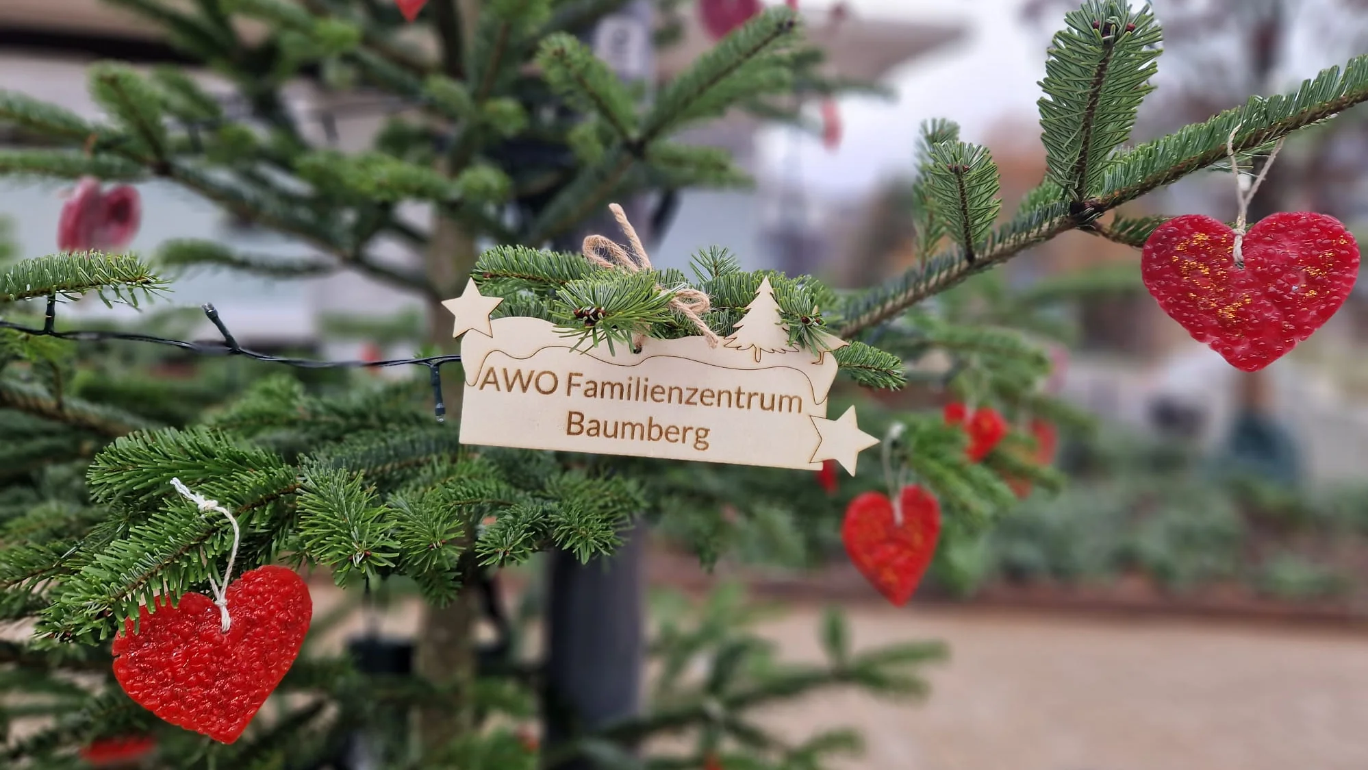 Das Bild zeigt eine Nahaufnahme eines Weihnachtsbaumzweiges, der geschmückt ist. Details: Dekoration: An den Zweigen hängen mehrere leuchtend rote, herzförmige Ornamente (vermutlich aus Wachs oder einem ähnlichen Material mit Glitzer), die an einer Schnur befestigt sind. Schild: Zentral im Bild ist ein kleines, helles Holzschild zu sehen, das an einem Tannenzweig hängt. Darauf ist in dunklen Buchstaben "AWO Familienzentrum Baumberg" eingraviert. Das Schild ist außerdem mit kleinen stern- und baumförmigen Ausschnitten verziert. Beleuchtung: Im Hintergrund verläuft eine Lichterkette mit kleinen Lichtern. Fokus: Der Fokus liegt auf dem Schild und den vorderen roten Herzen, während der Hintergrund unscharf gehalten ist.