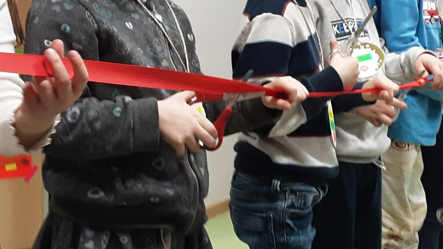 Auf dem Bild sieht man eine Gruppe von Kindern, die nebeneinander in einer Reihe stehen und gemeinsam ein rotes Band halten, als würden sie gleich eine Art „Banddurchschnitt“ oder Eröffnungszeremonie machen.