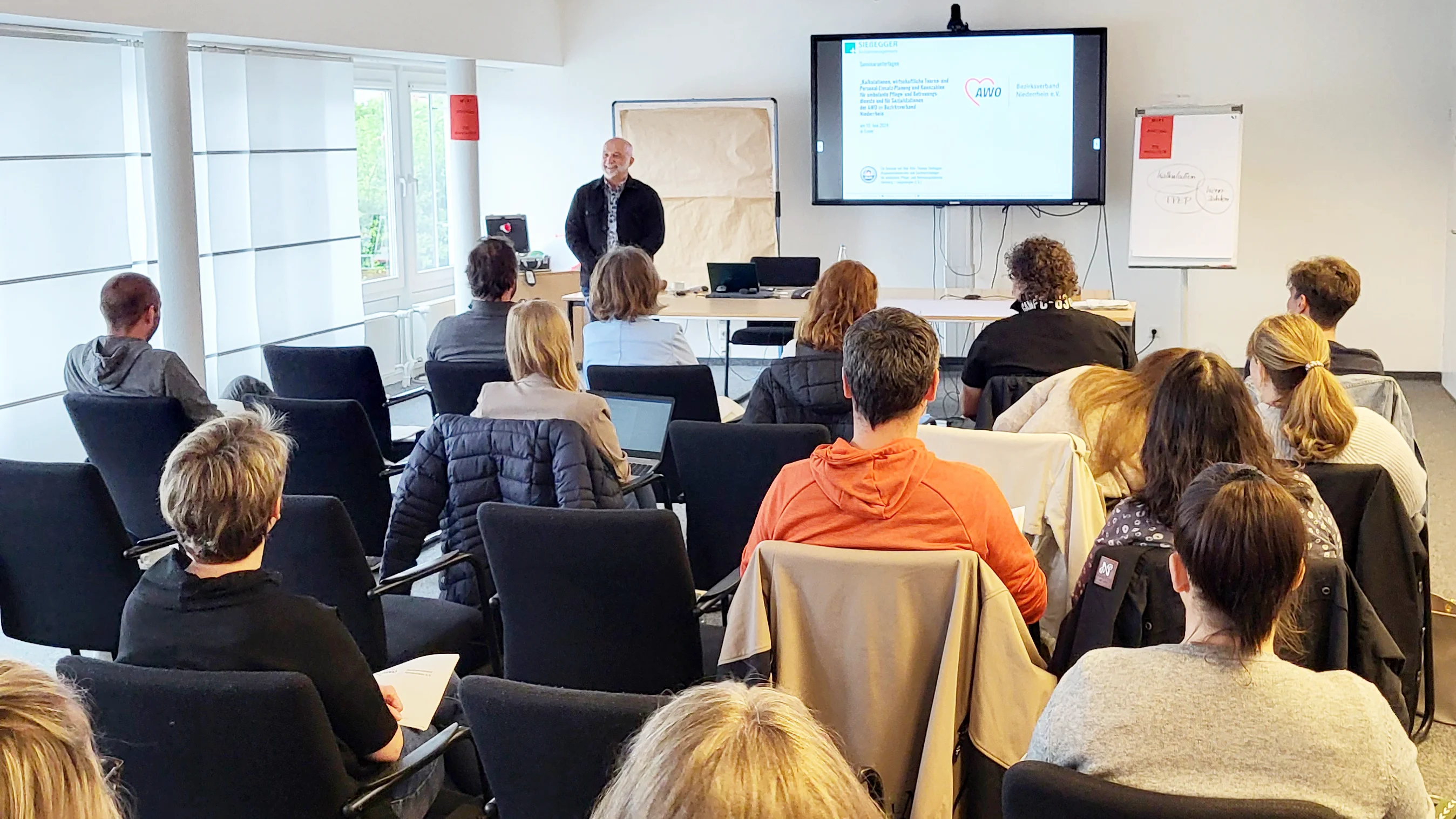 Das Foto zeigt einen Seminarraum, in dem Teilnehmende in Reihen sitzen und eine Präsentation an der Front des Raumes betrachten. Auf der Präsentationswand ist ein Teil einer PowerPoint-Folie zu sehen, die Text und Aufzählungspunkte enthält. Leider ist der genaue Inhalt von dieser Perspektive aus nicht klar erkennbar. Der Raum hat weiße Wände, und auf einer Seite befinden sich Fenster, die Tageslicht hereinlassen. 