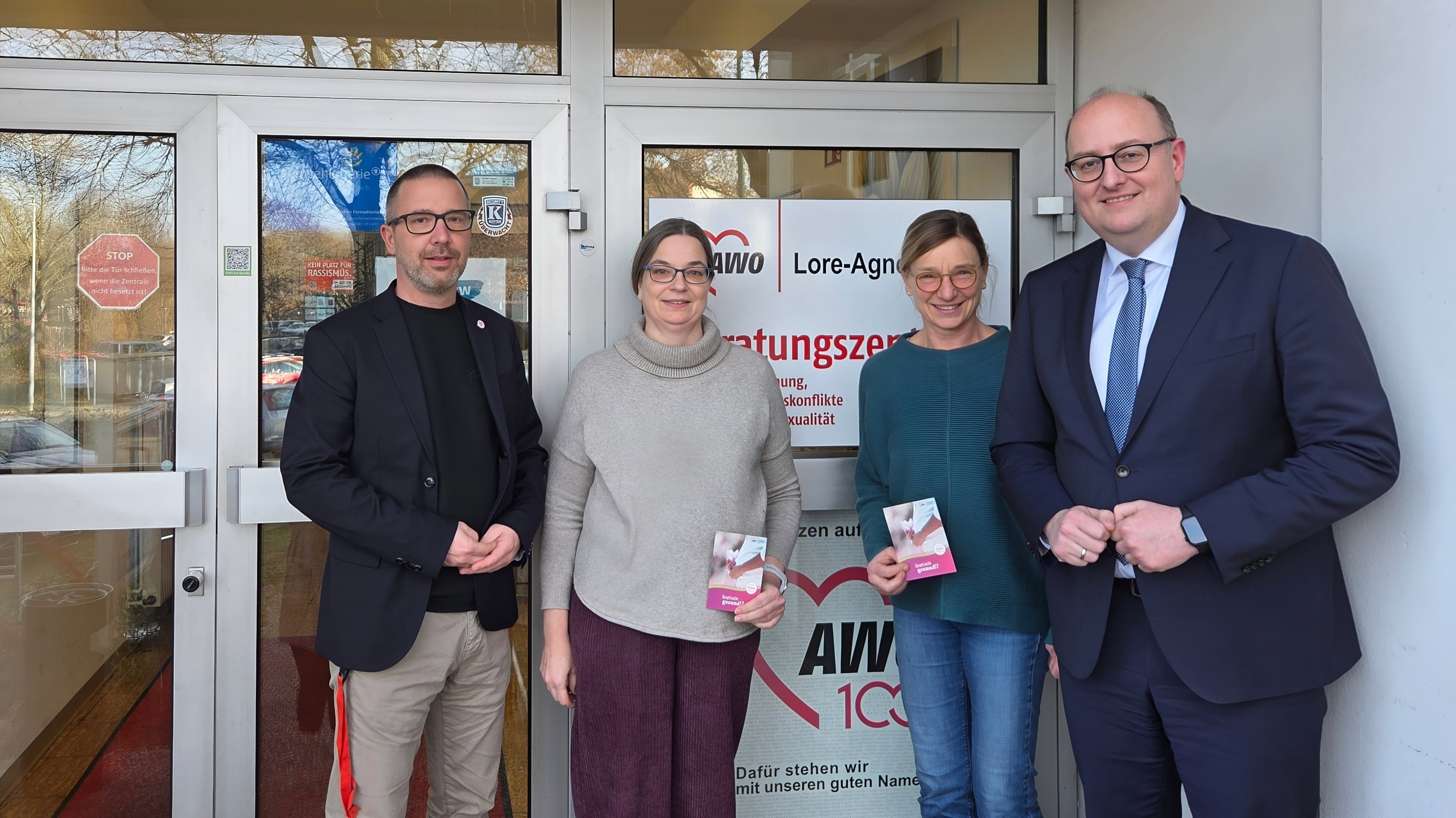 Auf dem Bild stehen vier erwachsene Personen vor einem Eingangsbereich mit Glastüren. Hinter ihnen ist ein Schild mit dem Logo der AWO und der Bezeichnung „Lore-Agnes-… Beratungszentrum“ zu sehen, außerdem weitere Texte zu Beratungsthemen. Die Personen halten Informationsflyer in der Hand und wirken wie zu einem offiziellen oder beruflichen Anlass fotografiert, etwa bei einem Besuch oder Termin vor dem Beratungszentrum.