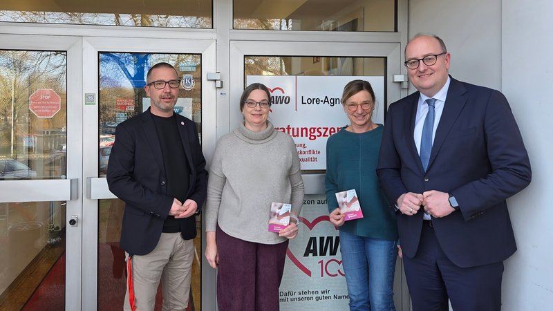 Auf dem Bild stehen vier erwachsene Personen vor einem Eingangsbereich mit Glastüren. Hinter ihnen ist ein Schild mit dem Logo der AWO und der Bezeichnung „Lore-Agnes-… Beratungszentrum“ zu sehen, außerdem weitere Texte zu Beratungsthemen. Die Personen halten Informationsflyer in der Hand und wirken wie zu einem offiziellen oder beruflichen Anlass fotografiert, etwa bei einem Besuch oder Termin vor dem Beratungszentrum.