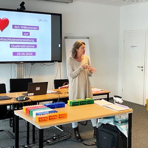 Das Foto zeigt Oda Bakuhn in dem Seminarraum. Im Hintergrund ist ein Whiteboard mit dem Text "Herzlich Willkommen zur Abschlussveranstaltung Qualimaßnahme 29.05.2024"