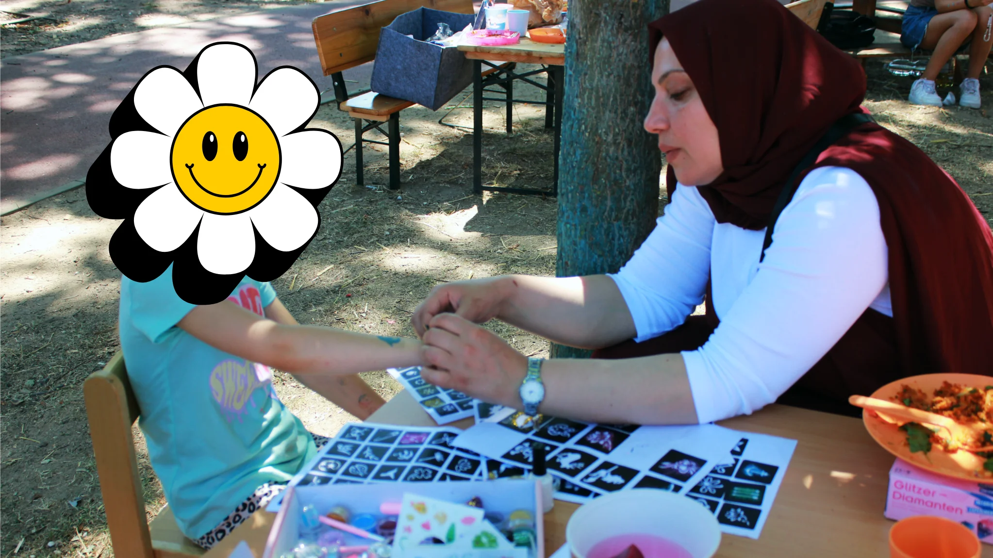 Im Bild sieht man eine Szene von einem sommerlichen Kinder- bzw. Familienfest im Freien. Eine erwachsene Person sitzt an einem Tisch und bringt einem Kind ein Glitzer‑Tattoo oder ähnlichen Körperschmuck auf den Arm an; auf dem Tisch liegen zahlreiche Schablonen und Bastelmaterialien, außerdem Becher und Schalen mit Essen bzw. Getränken.
