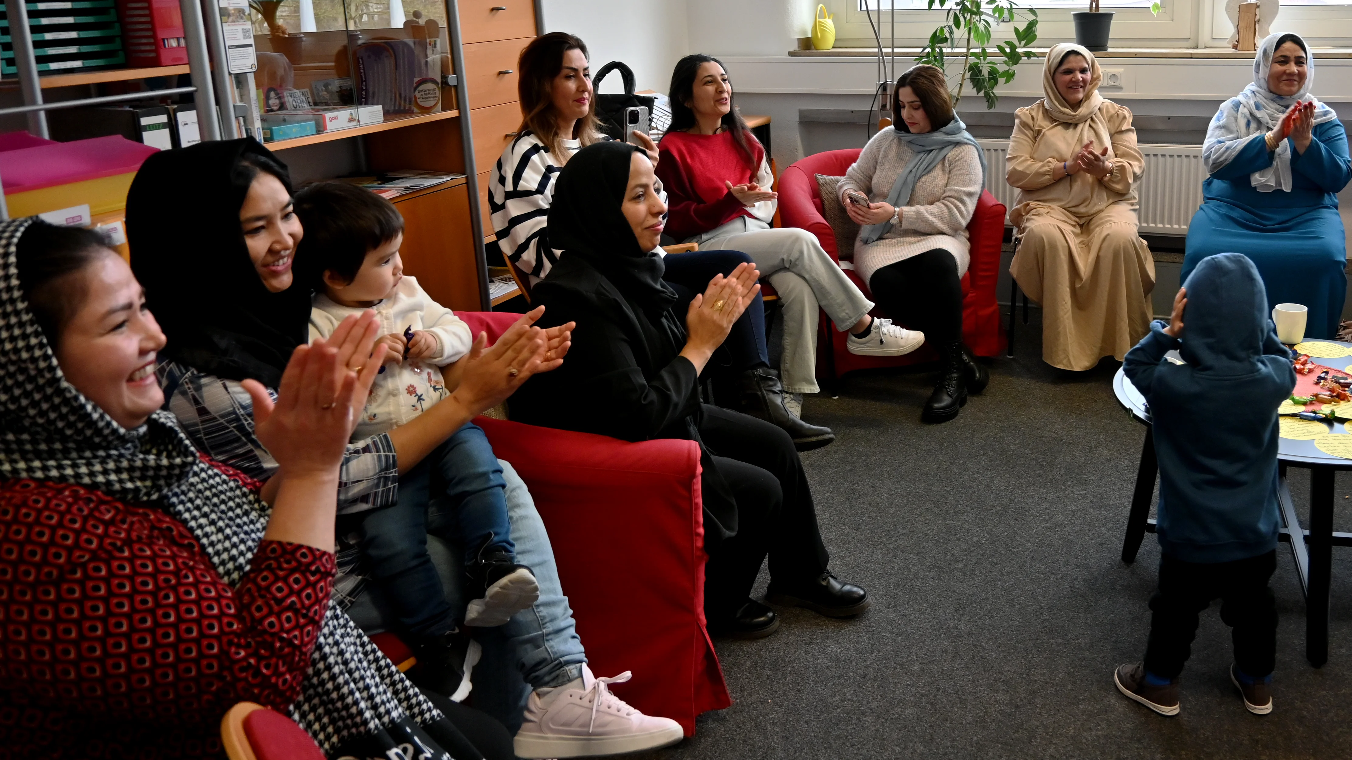 Das Foto zeigt eine Gruppe von Frauen, die in einem Raum auf roten Sesseln und Stühlen sitzen. Sie klatschen und lächeln, was darauf hindeutet, dass sie an einer fröhlichen oder feierlichen Aktivität teilnehmen. Einige Frauen tragen Kopftücher, andere haben offene Haare. In der Mitte des Bildes hält eine Frau ein kleines Kind auf ihrem Schoß, während vorne ein weiteres Kind in einem blauen Kapuzenpullover steht und sich möglicherweise die Ohren zuhält. Im Hintergrund sieht man Regale mit Ordnern und Büchern, sowie Pflanzen und Fenster, die den Raum erhellen.